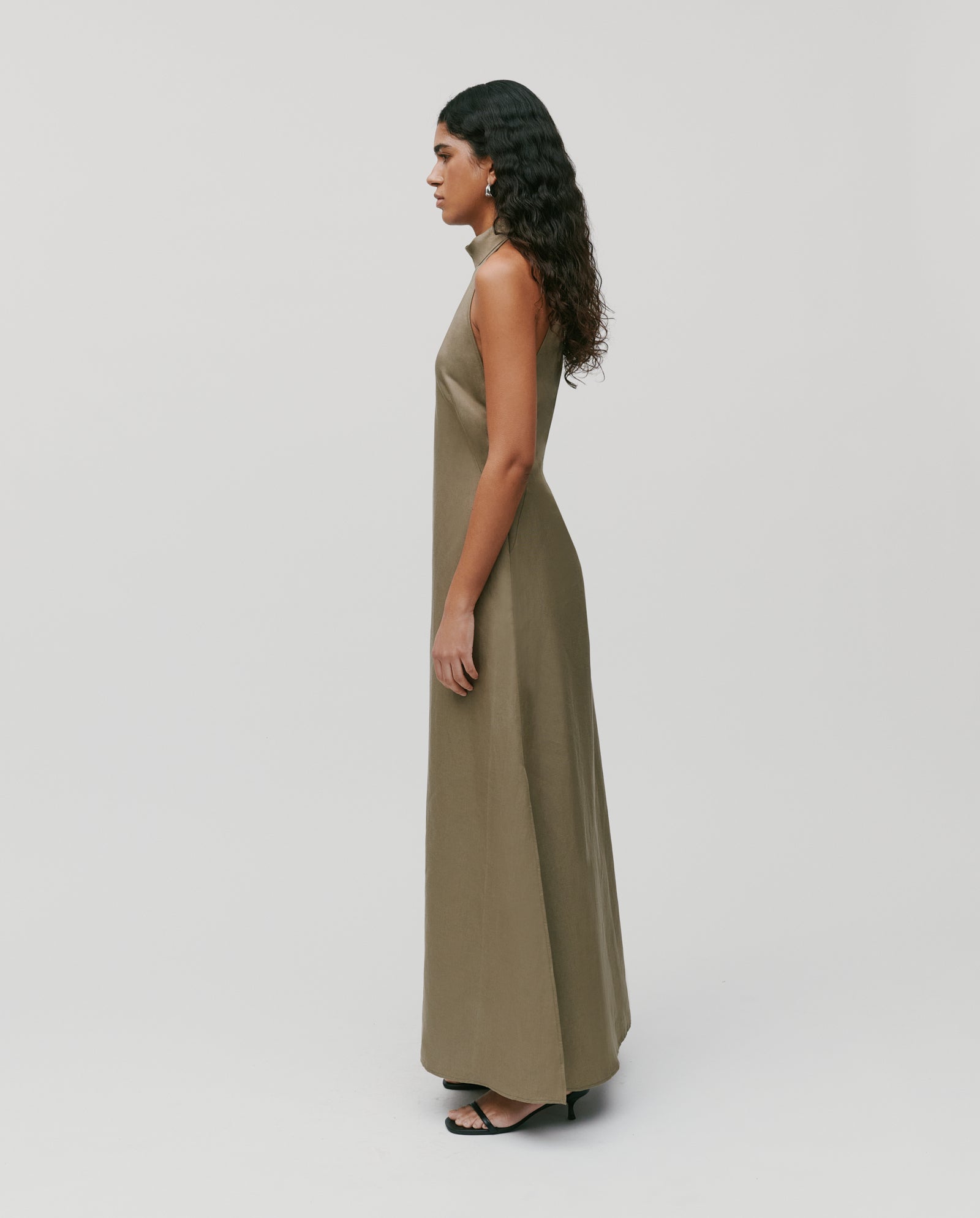 A woman in profile wears the IVY OAK NELA Dress, a sleeveless olive-green maxi, paired with black heels against a plain background.