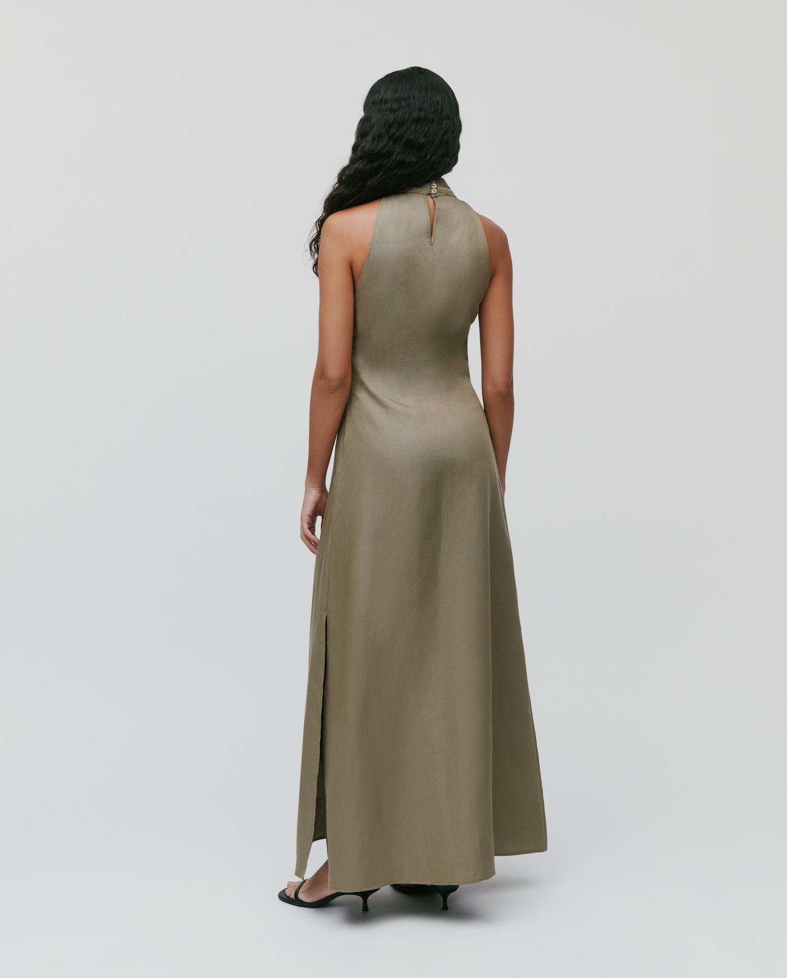 A woman stands facing away from the camera in the NELA Dress by IVY OAK, an olive green linen blend maxi with a neckholder design and side slit.