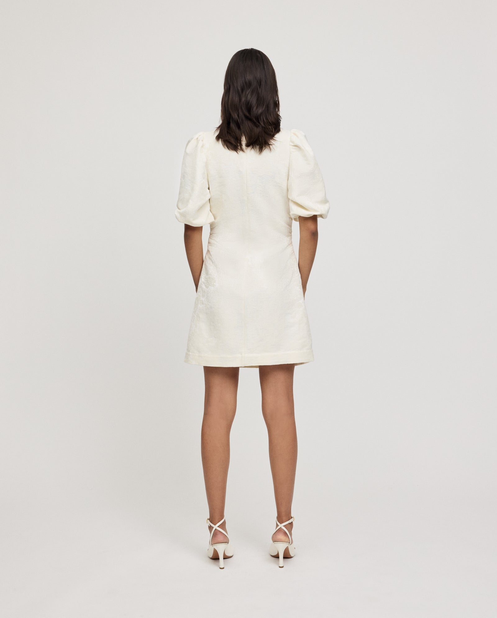 A woman stands with her back to the camera, showcasing the back zipper of the IVY OAK NISSA Dress—a jacquard mini dress with puff sleeves—against a plain background, paired with heels.