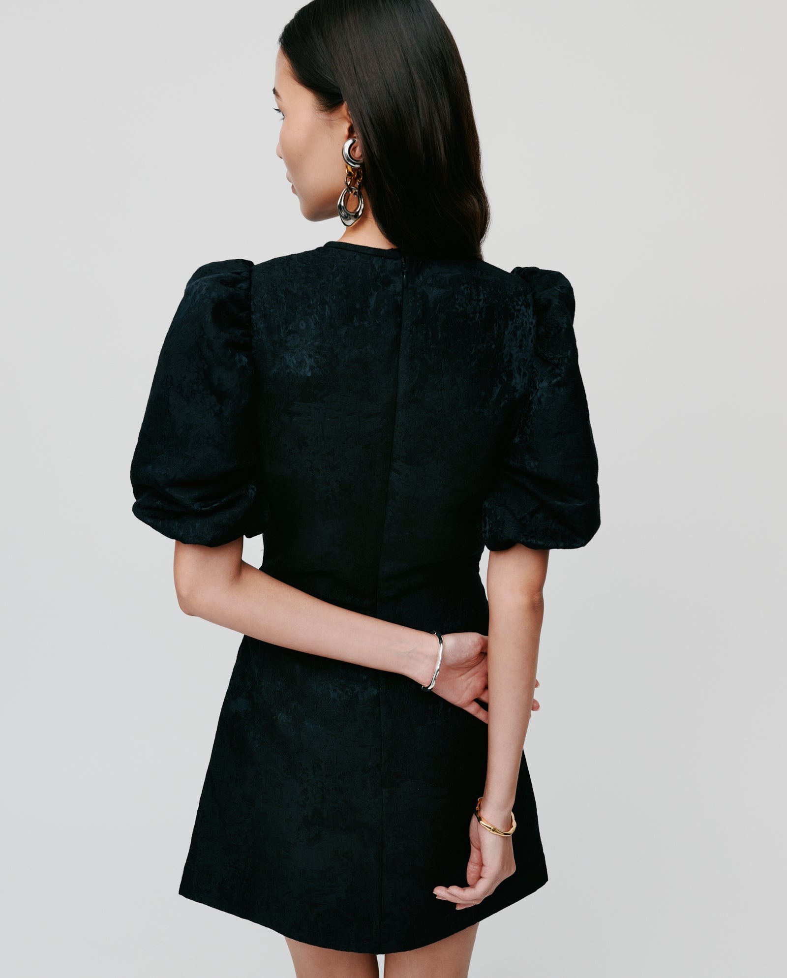 A woman in the IVY OAK NISSA Dress, a black jacquard mini with subtle puff sleeves, stands with her arms behind her back, facing away from the camera.