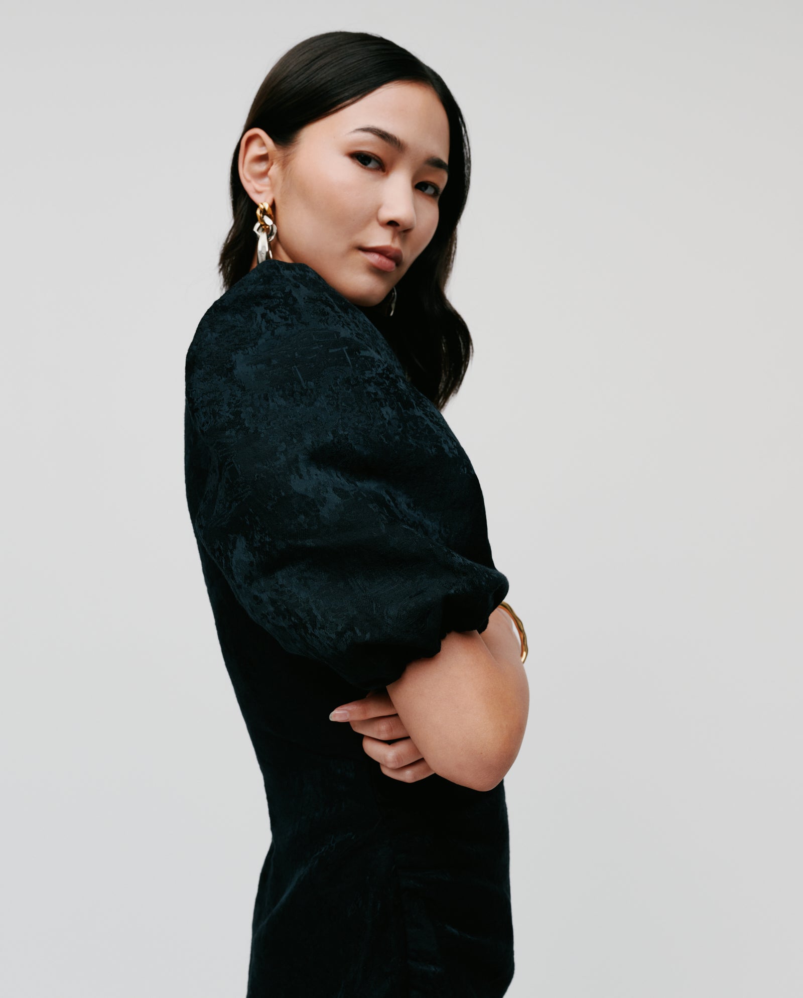 Woman with long dark hair in a black NISSA Dress by IVY OAK with subtle puff sleeves, posing confidently, arms crossed against a plain background.