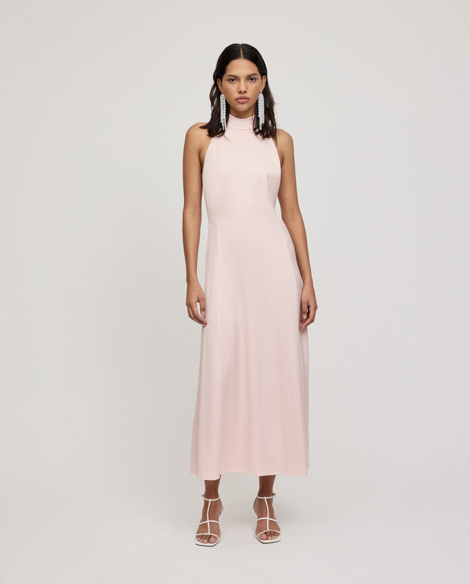 Woman in a sleeveless, light pink IVY OAK DARCI ANN dress with an American shoulder design and white strappy heels, standing against a plain backdrop.