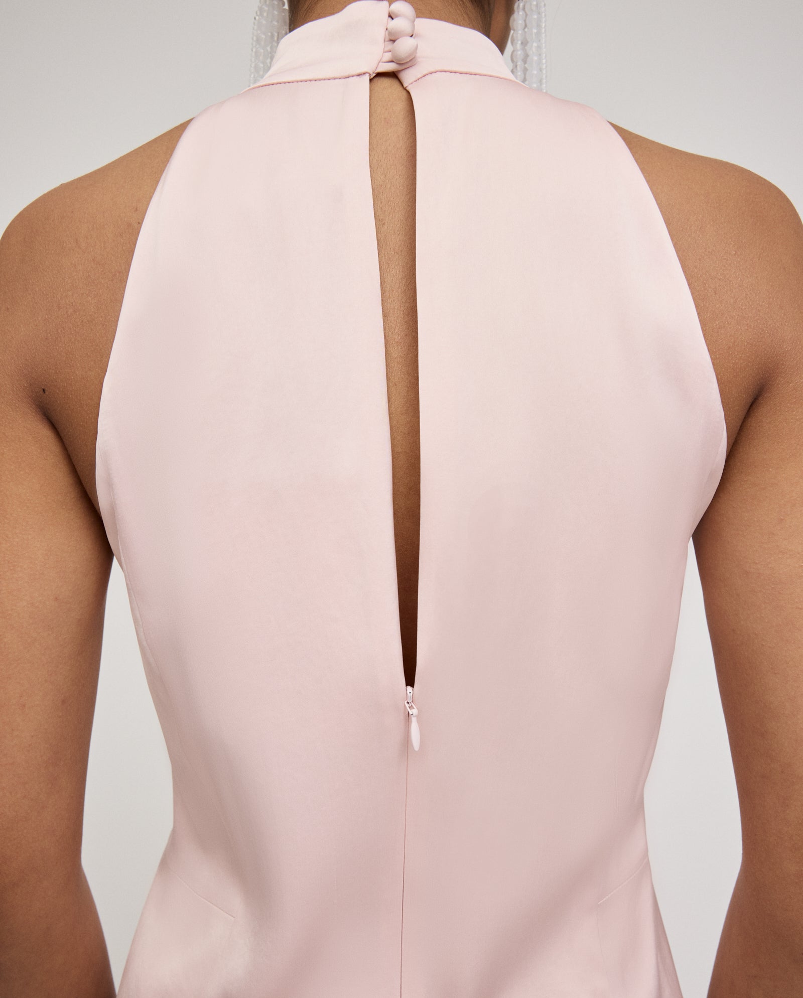 Back view of a person wearing the IVY OAK DARCI ANN Dress, a pale pink midi halter dress featuring a back slit and button closure at the neck.