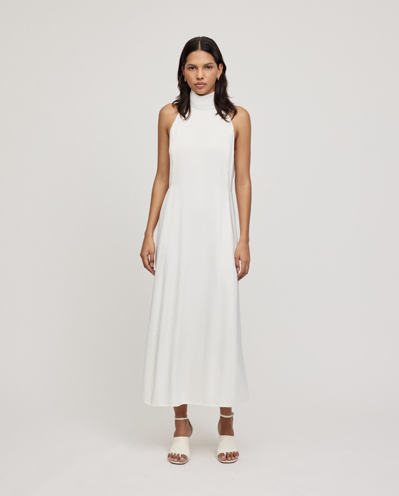 A woman in the IVY OAK DARCI ANN sleeveless, high-neck American shoulder dress with a back zipper and white sandals stands against a plain background.