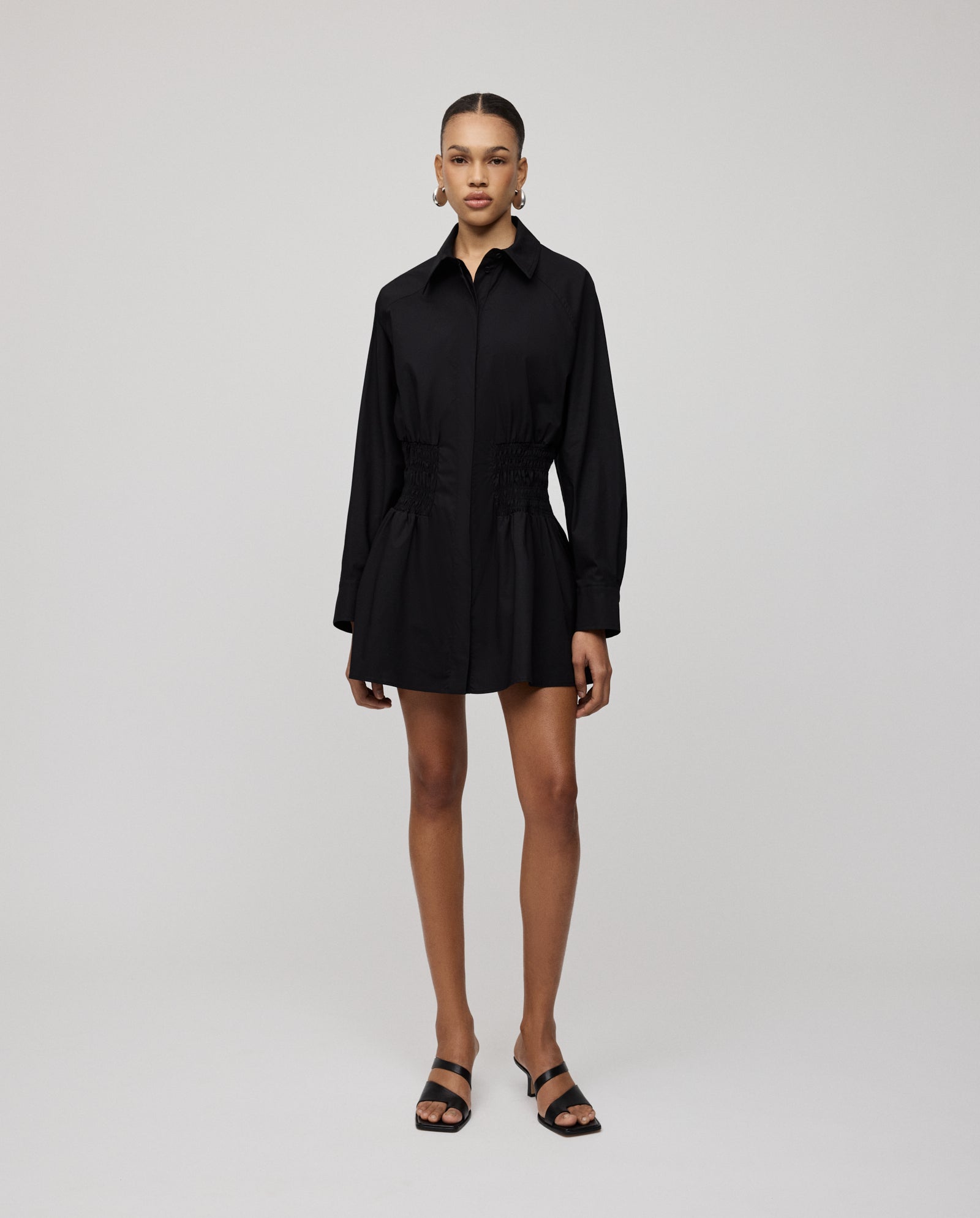 A woman stands against a plain background wearing the IVY OAK MARYNA Dress—a black, long-sleeve mini shirt dress with a ruched waist and button placket—paired with black sandals.
