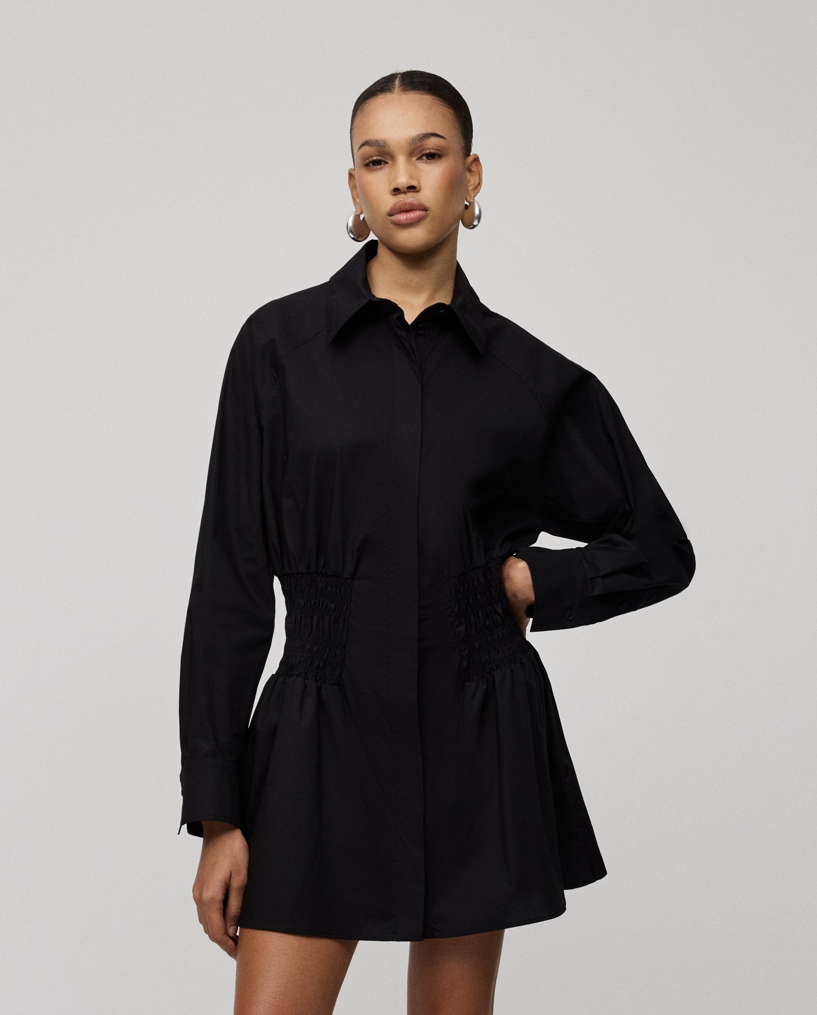 A woman wears the IVY OAK MARYNA Dress, a long-sleeve black mini shirt dress with a button placket and gathered waist, standing against a plain background.