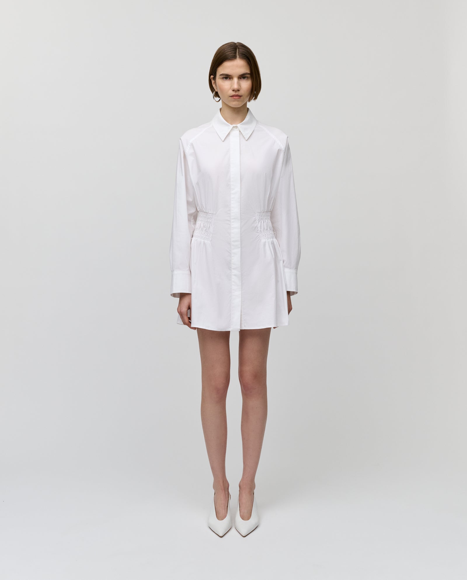 A woman stands against a white background wearing the IVY OAK MARYNA Dress, a long-sleeved mini shirt dress with smock detail, elegantly paired with white heels.