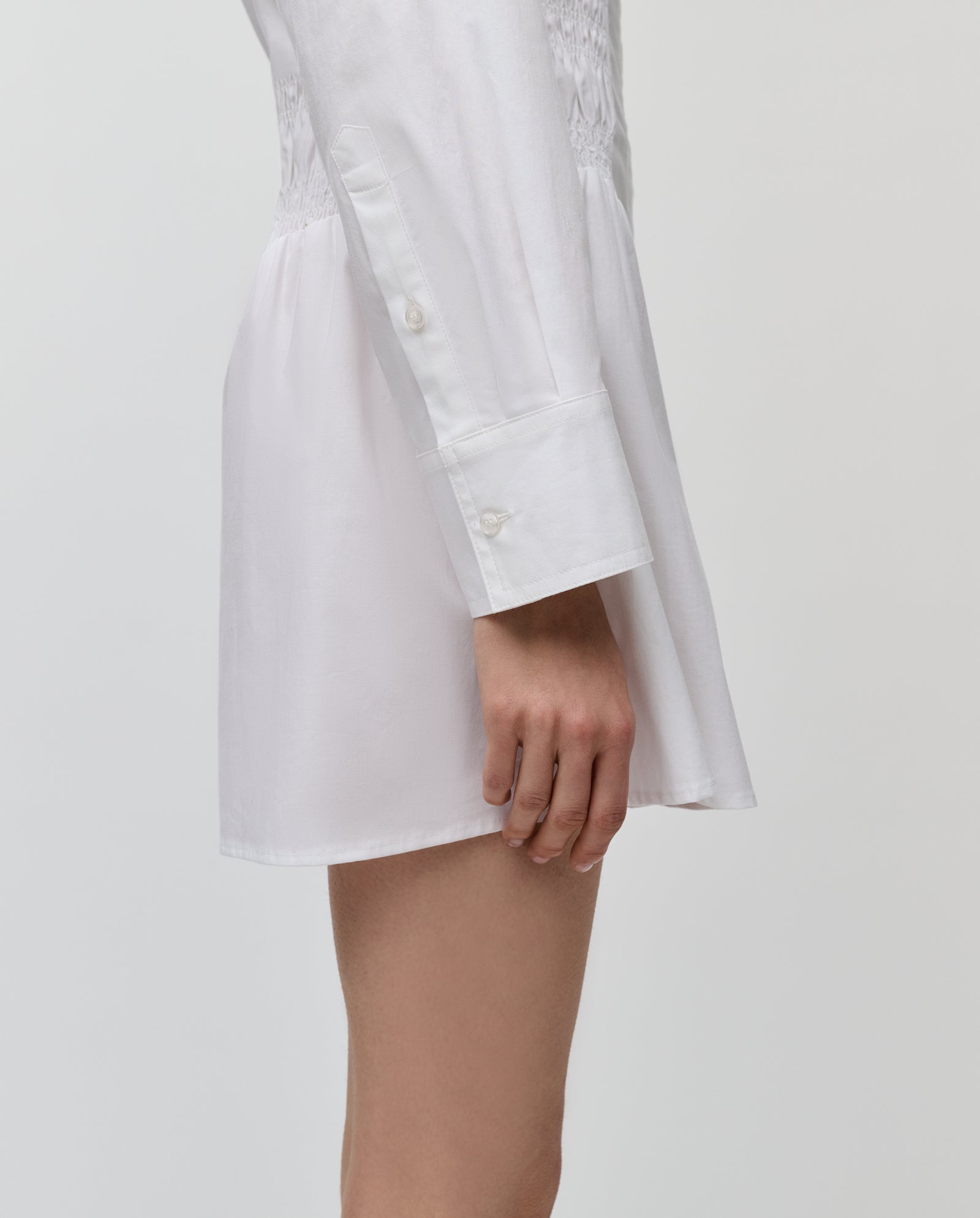 The IVY OAK MARYNA Dress is a long-sleeved white mini shirt dress, shown in profile with the wearer’s arm relaxed.