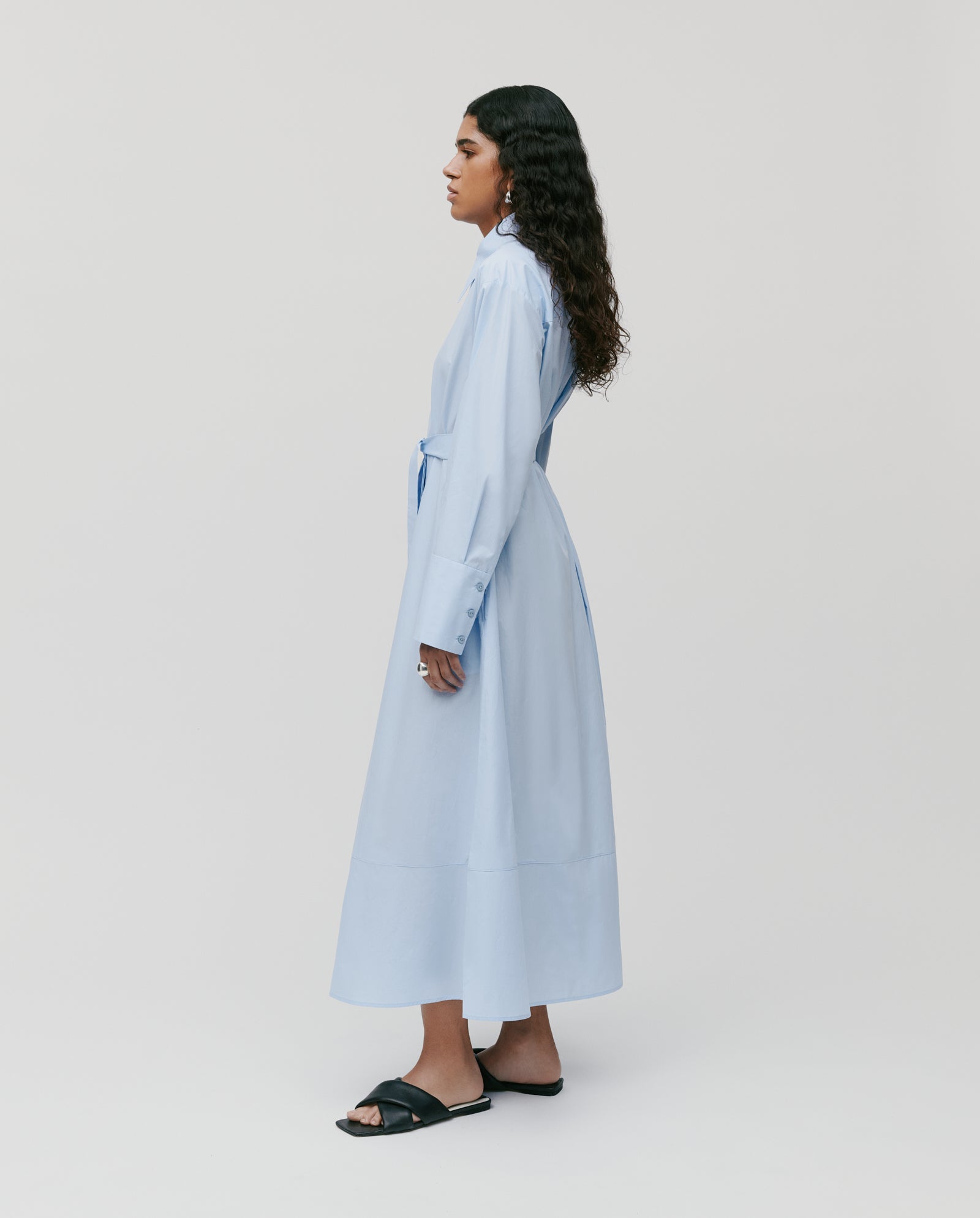 A person stands sideways against a plain background wearing the IVY OAK DINA ROSA light blue, long-sleeved cotton summer dress with adjustable cuffs and black sandals.