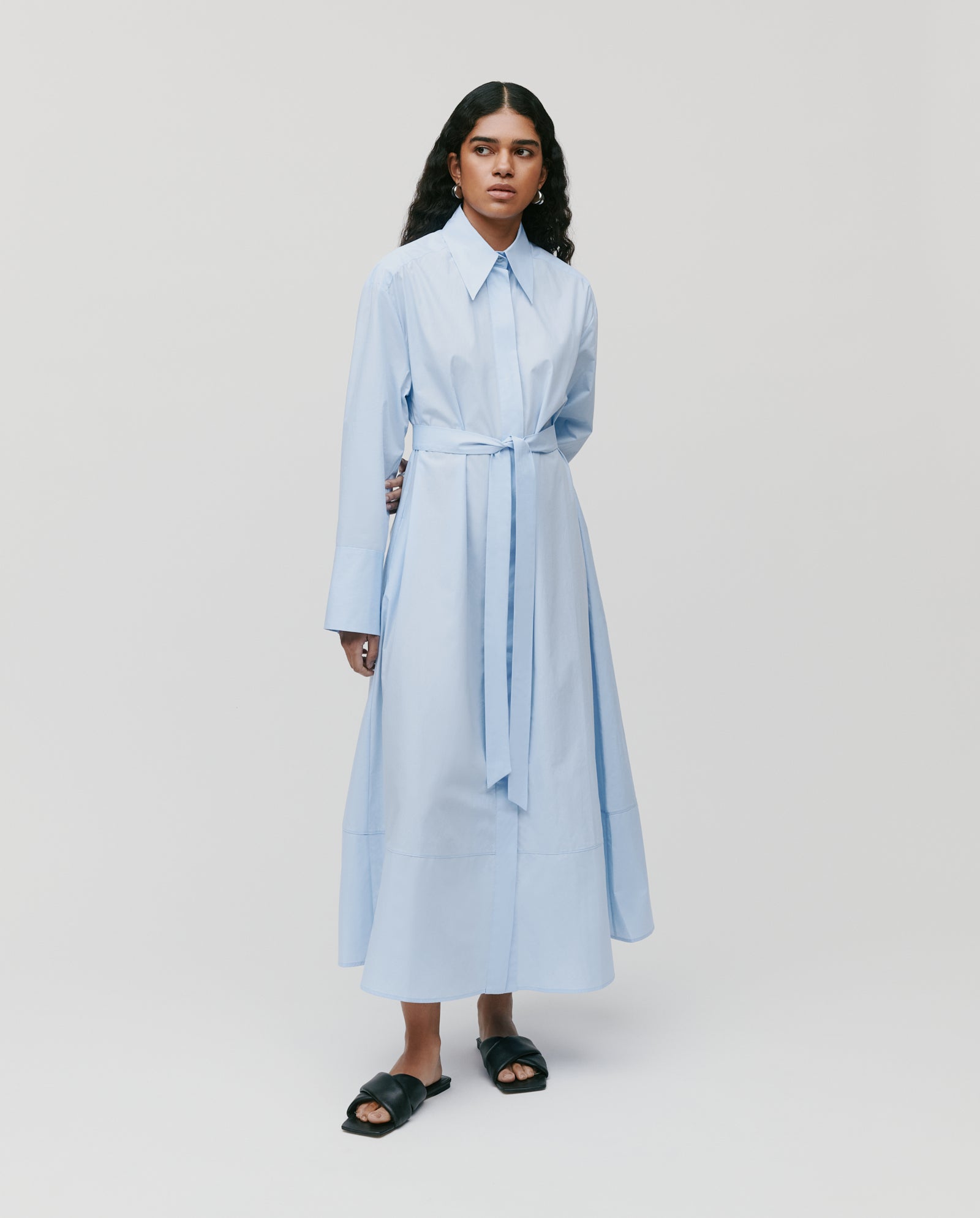 A woman stands against a plain background wearing the IVY OAK DINA ROSA Dress, a long light blue cotton summer dress with a waist tie, classic shirt design, and adjustable cuffs, paired with black slide sandals.