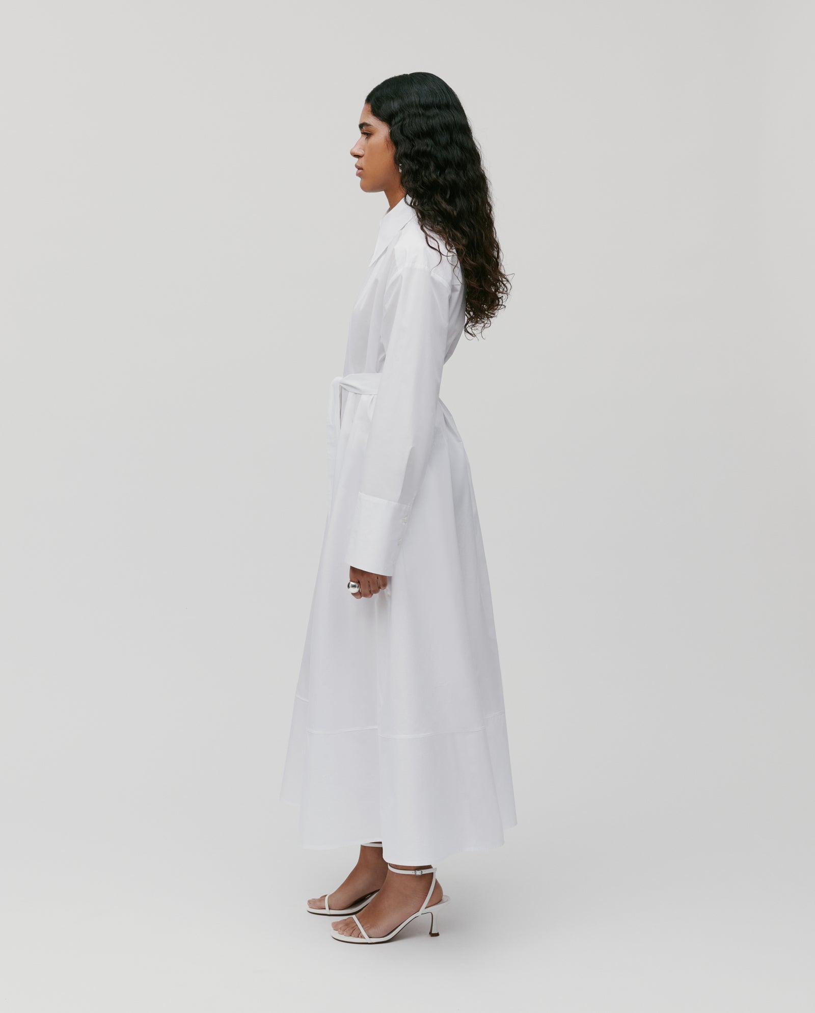 A woman wearing the IVY OAK DINA ROSA long white cotton summer dress with adjustable cuffs and white heels, standing in profile against a plain light background.