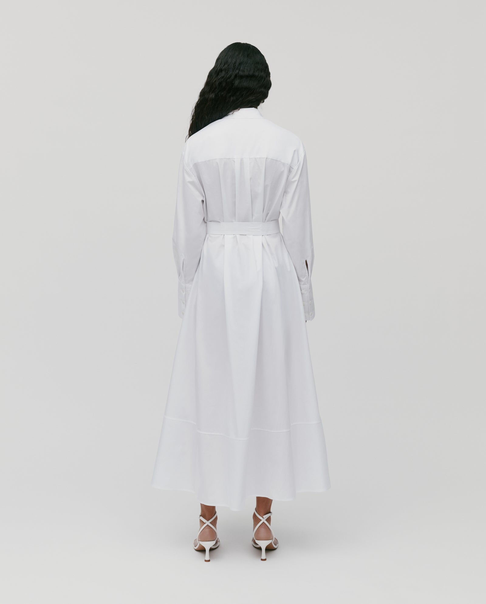 A woman in the IVY OAK DINA ROSA Dress, a long white cotton summer piece with adjustable cuffs, stands with her back to the camera against a plain background, styled elegantly with matching heels.