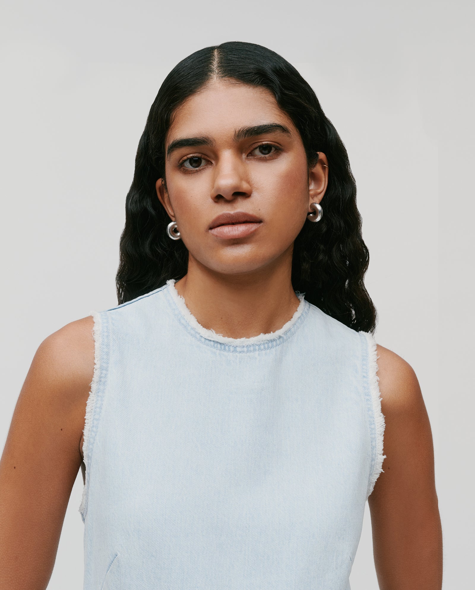 Woman with long wavy hair wears the IVY OAK MARCA ANN Denim Dress, a sleeveless, ankle-length piece featuring subtle fringe details, paired with silver hoop earrings, shown facing forward.