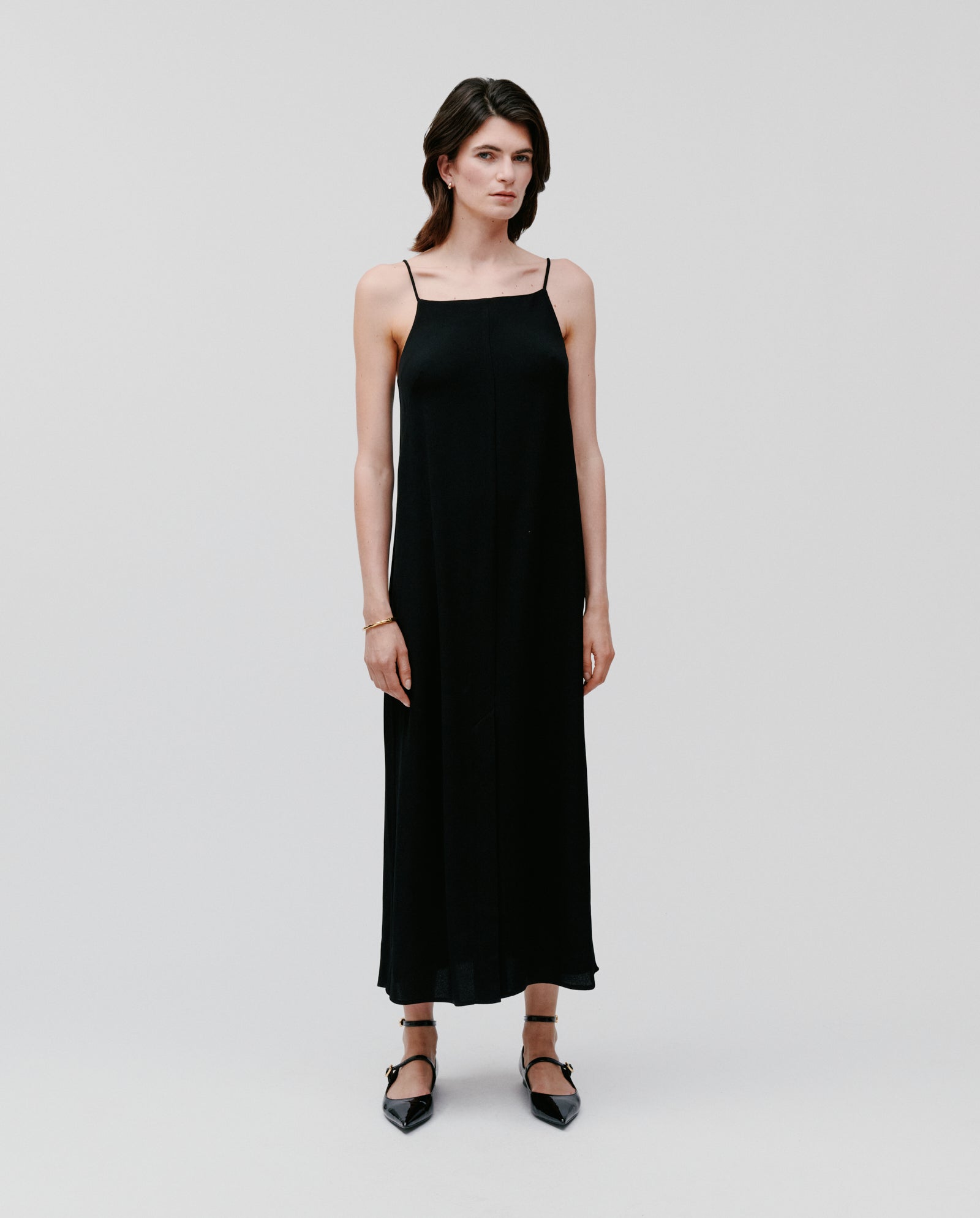A woman wears the IVY OAK NAVA Dress—a sleeveless black crepe maxi styled with black pointed-toe flats—posing against a plain white background, perfect as a warm-weather essential.