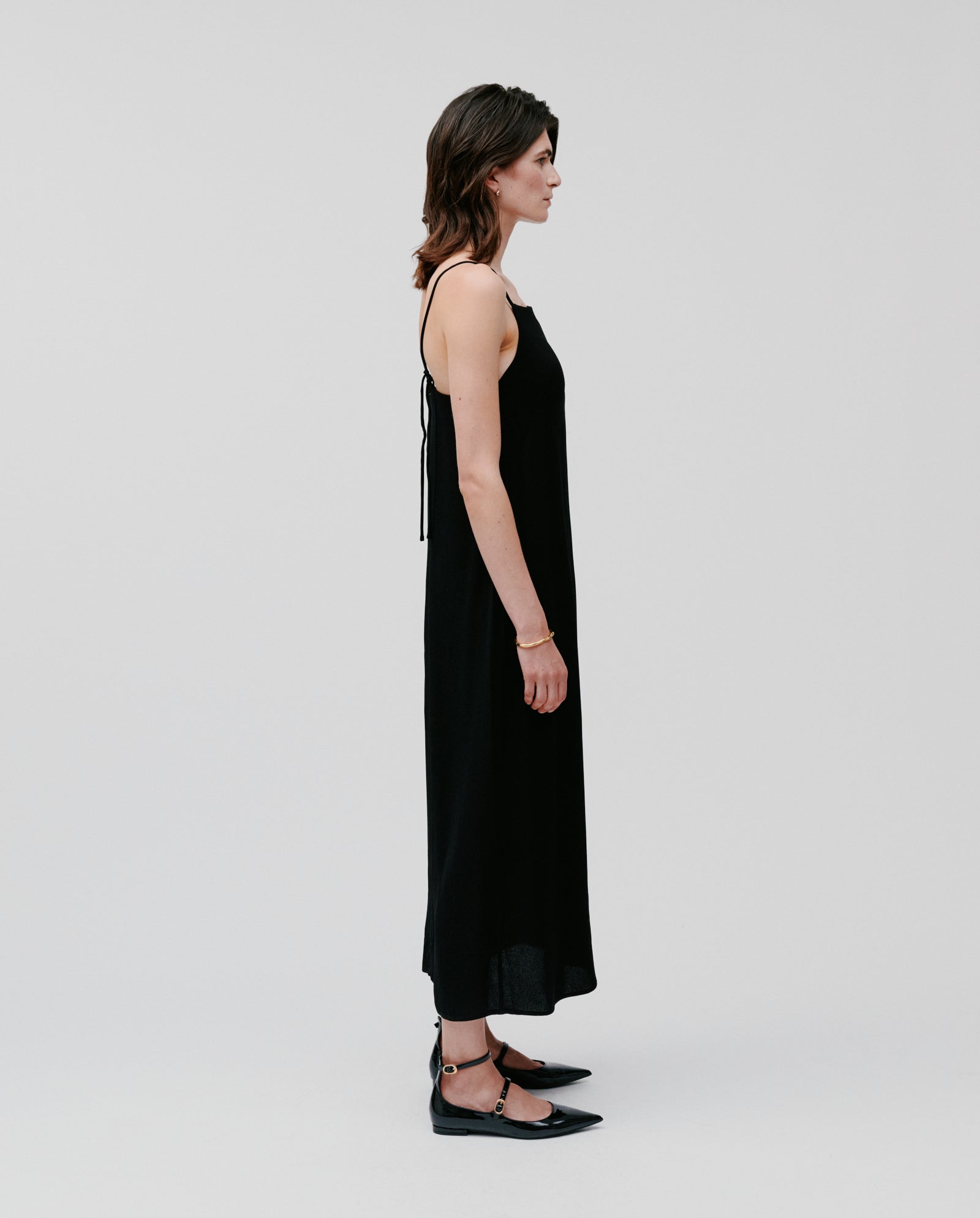 A woman in the IVY OAK NAVA Dress—sleeveless, black crepe fabric—pairs it with black flats while standing in profile against a white backdrop. This lightweight piece is a summer and warm-weather essential.