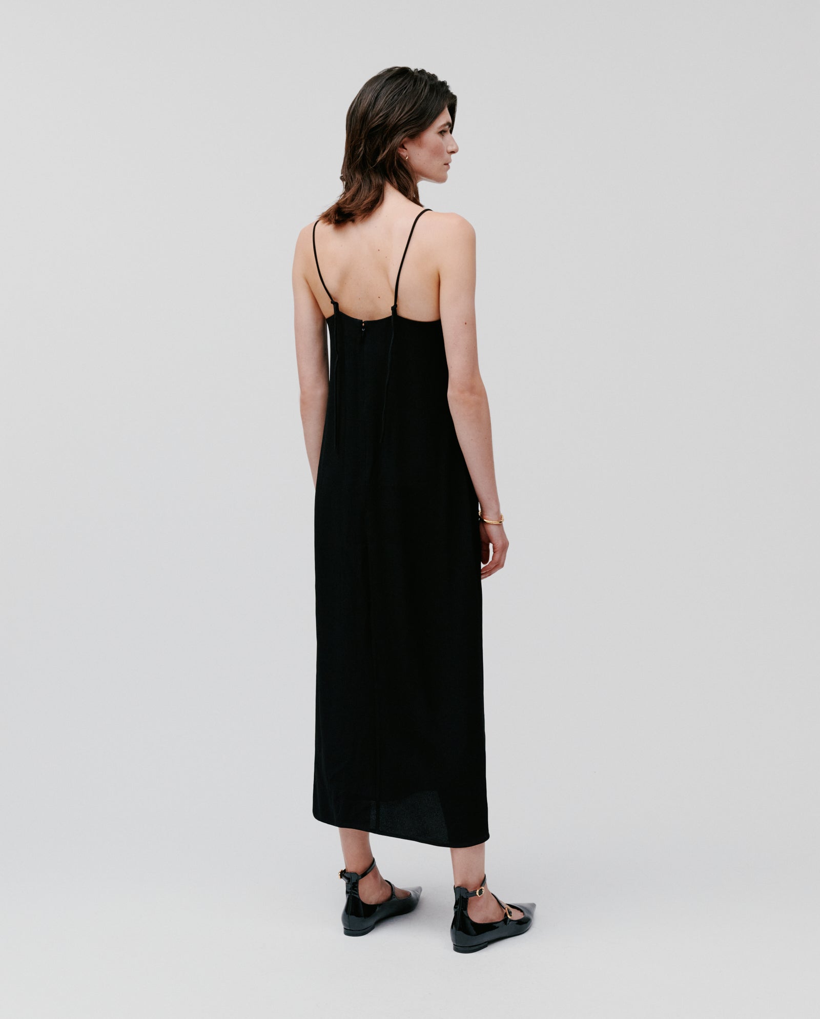 A woman faces away, wearing the IVY OAK NAVA Dress—a long black spaghetti strap lightweight summer dress—paired with black sandals against a plain background.