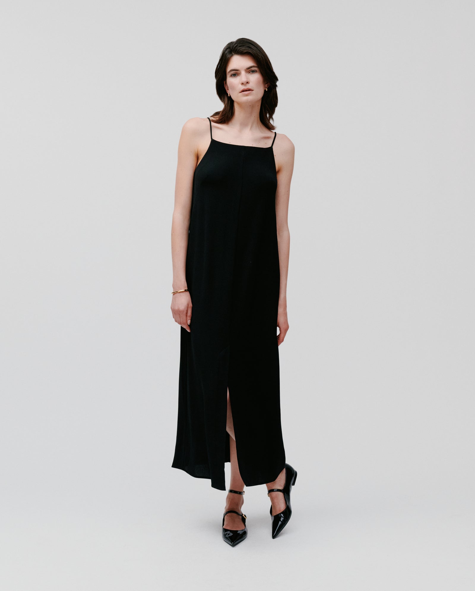 A woman wears the NAVA Dress by IVY OAK—a sleeveless black crepe midi dress with thin straps—paired with black heels against a plain background, creating an effortless warm-weather look.