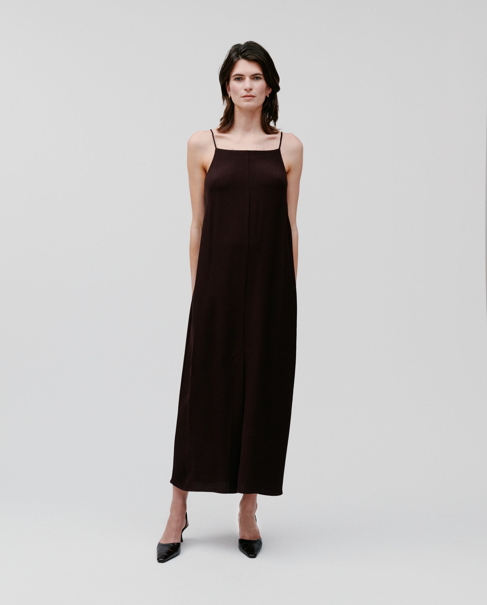 A woman models the IVY OAK NAVA Dress, a black sleeveless lightweight summer style, paired with black heels against a plain white background.