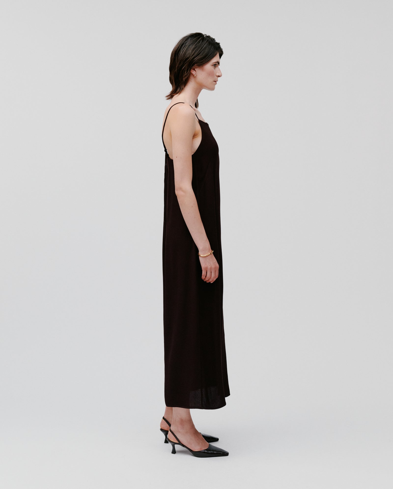A person stands in profile against a plain light background wearing the NAVA Dress by IVY OAK, a long black lightweight summer dress paired with black heels.