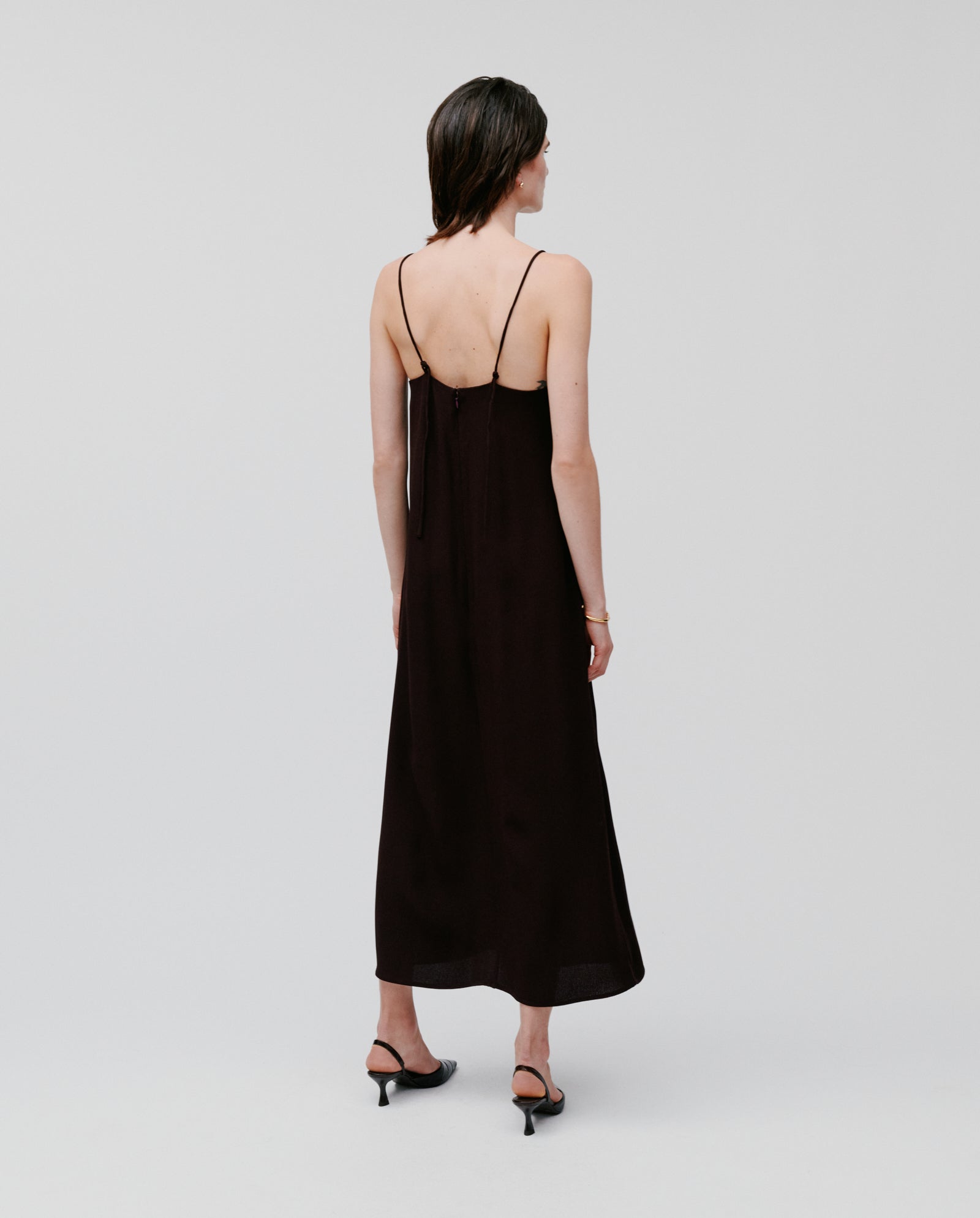 A woman in the IVY OAK NAVA Dress—black, backless, sleeveless, and lightweight—stands against a plain light background in heels. This summer dress is the perfect warm-weather essential for effortless style.