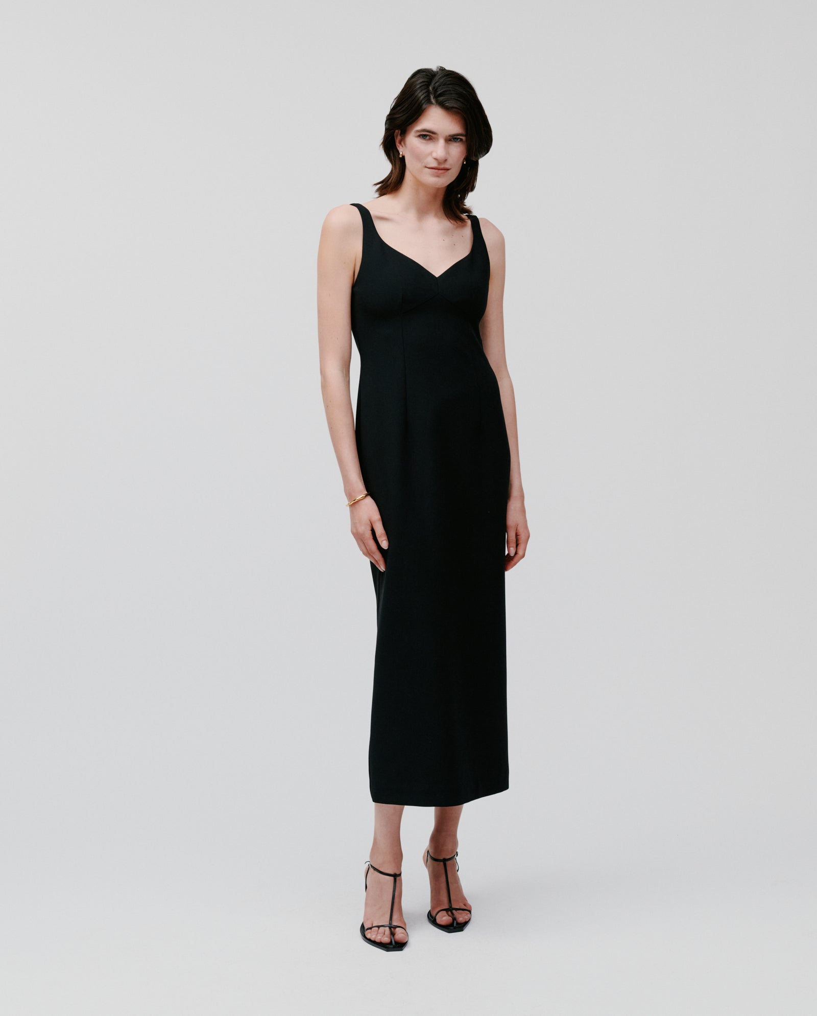 A woman wearing the IVY OAK NEPHELIA Dress, a sleeveless, ankle-length design in matte fabric, with heels, stands against a plain light background.