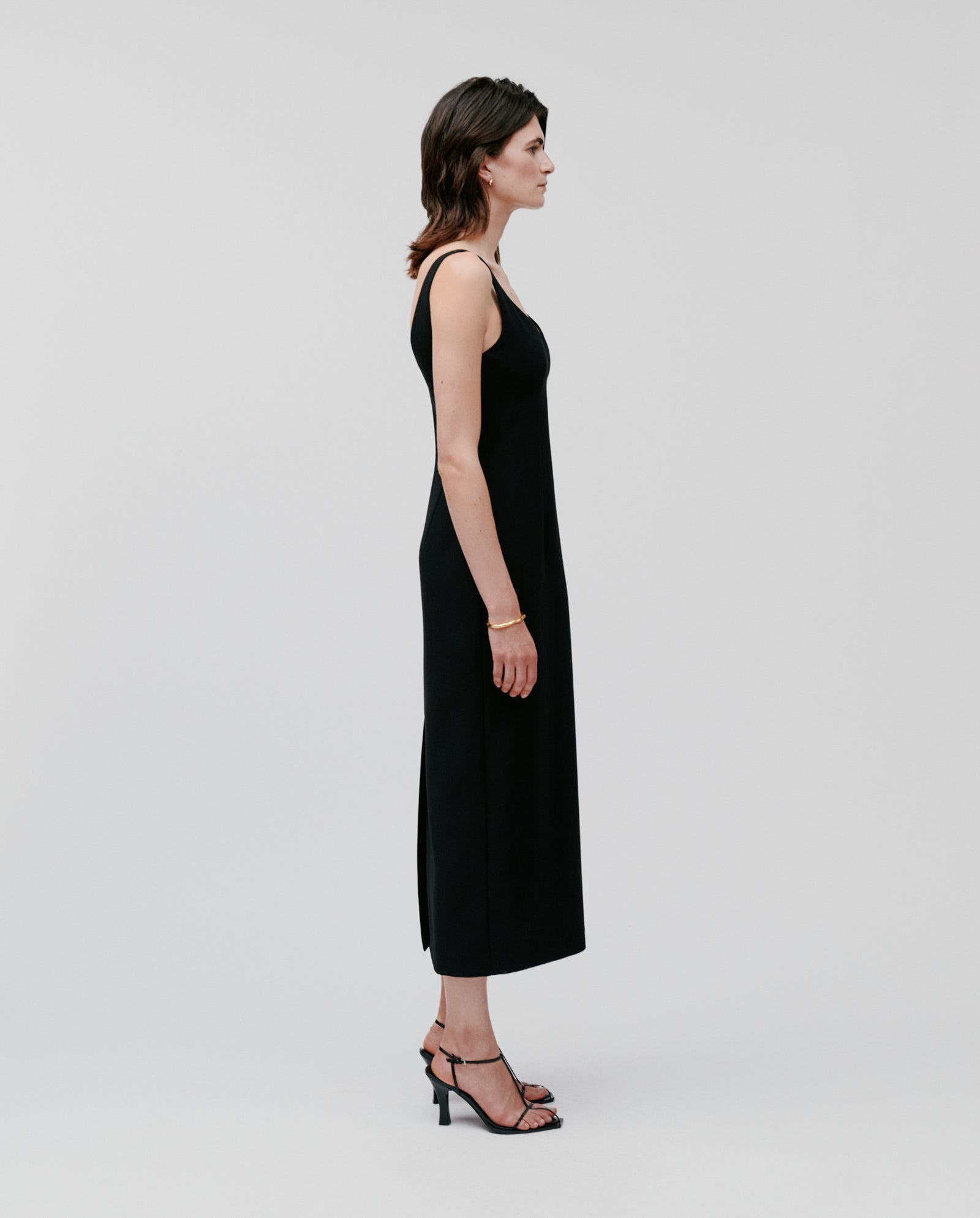 A woman wearing the IVY OAK NEPHELIA Dress, featuring a sleeveless matte finish, stands in profile against a plain white background with heels.