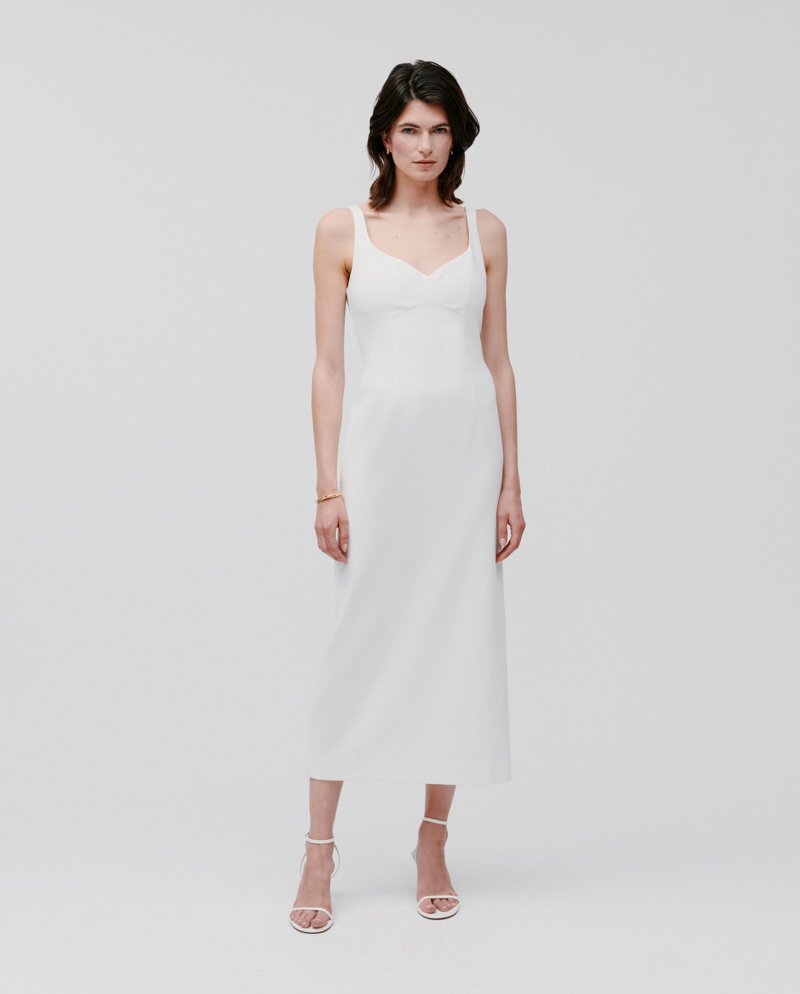 A woman wearing the IVY OAK NEPHELIA Dress—an ankle-length, matte white dress with a slit back—and sandals, standing against a plain white background.