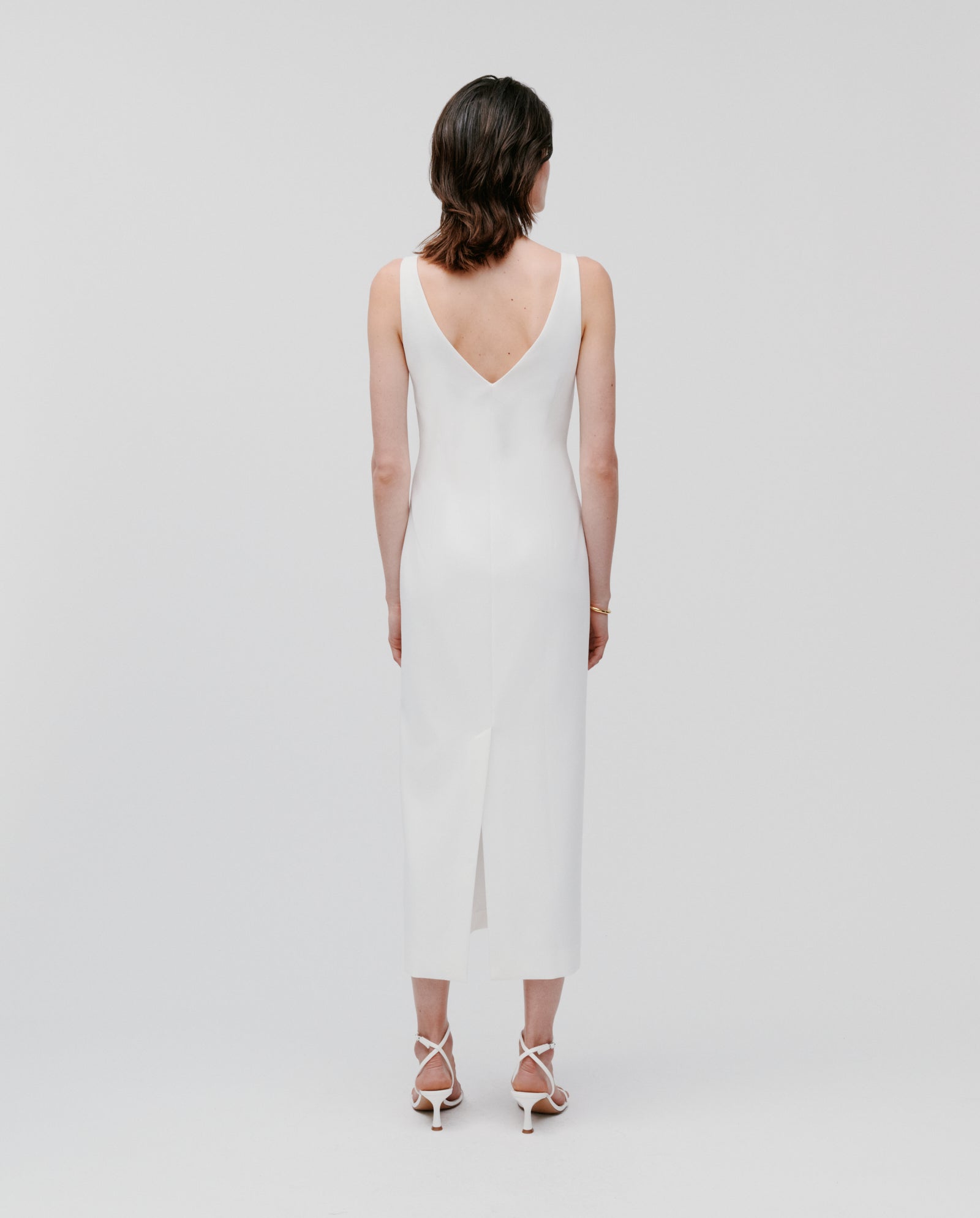 Woman models the IVY OAK NEPHELIA Dress, an ankle-length white dress with a slit back, paired with heels and photographed from behind against a plain background.