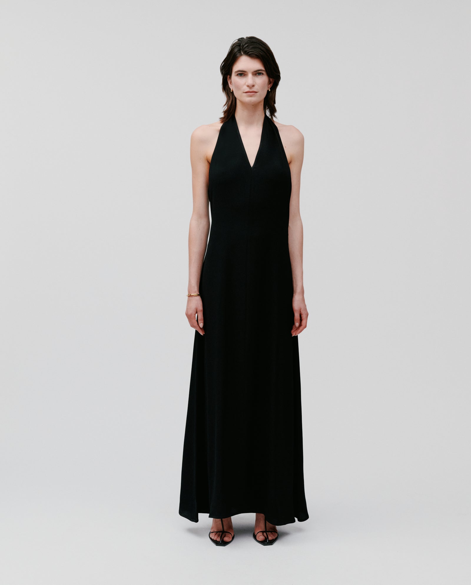 A person wears the NESTA Dress by IVY OAK, a sleeveless black neckholder style in crepe fabric, paired with black heels and standing against a plain light background.