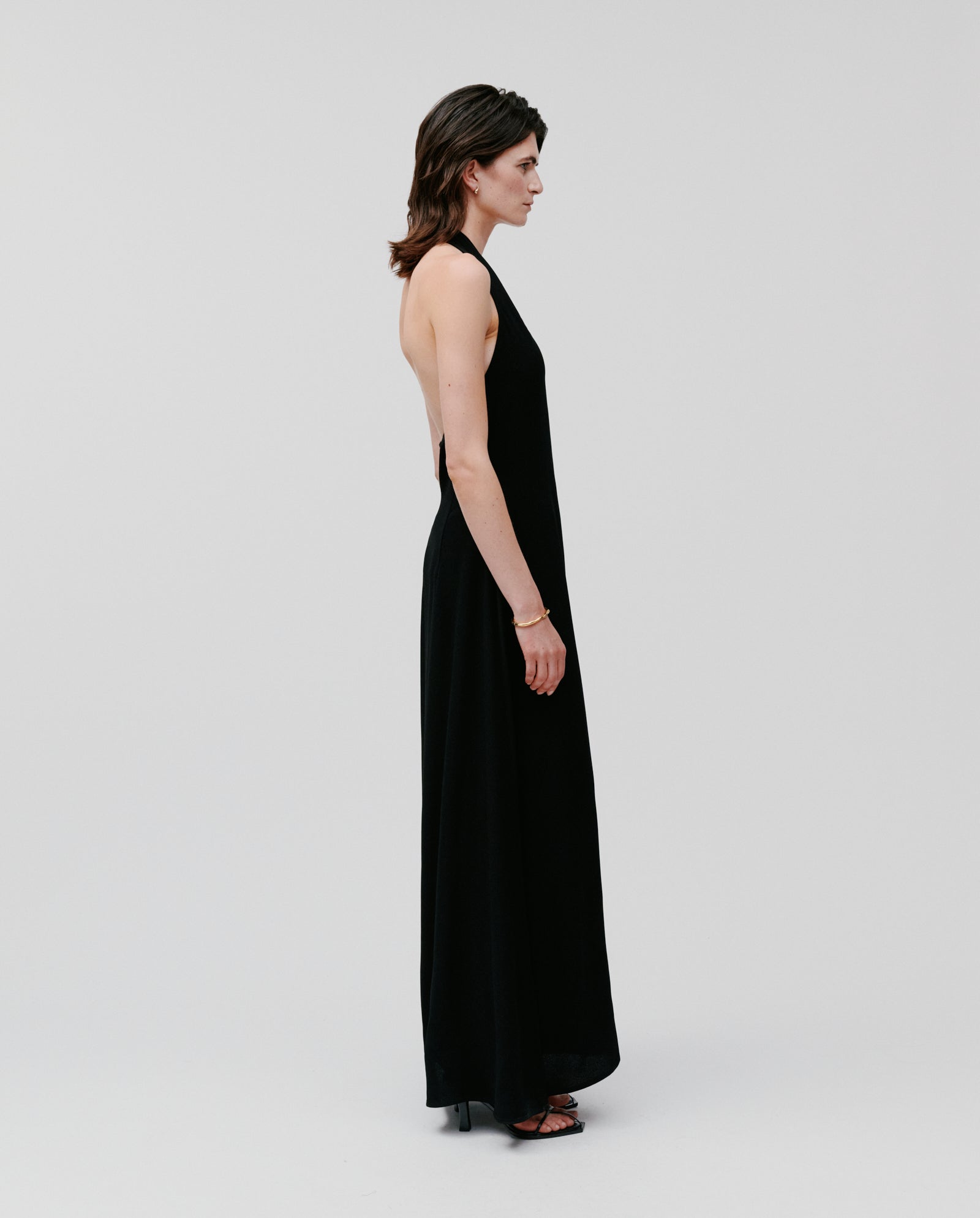 Woman wearing the IVY OAK NESTA sleeveless black halterneck crepe dress with heels, standing sideways against a plain light background.