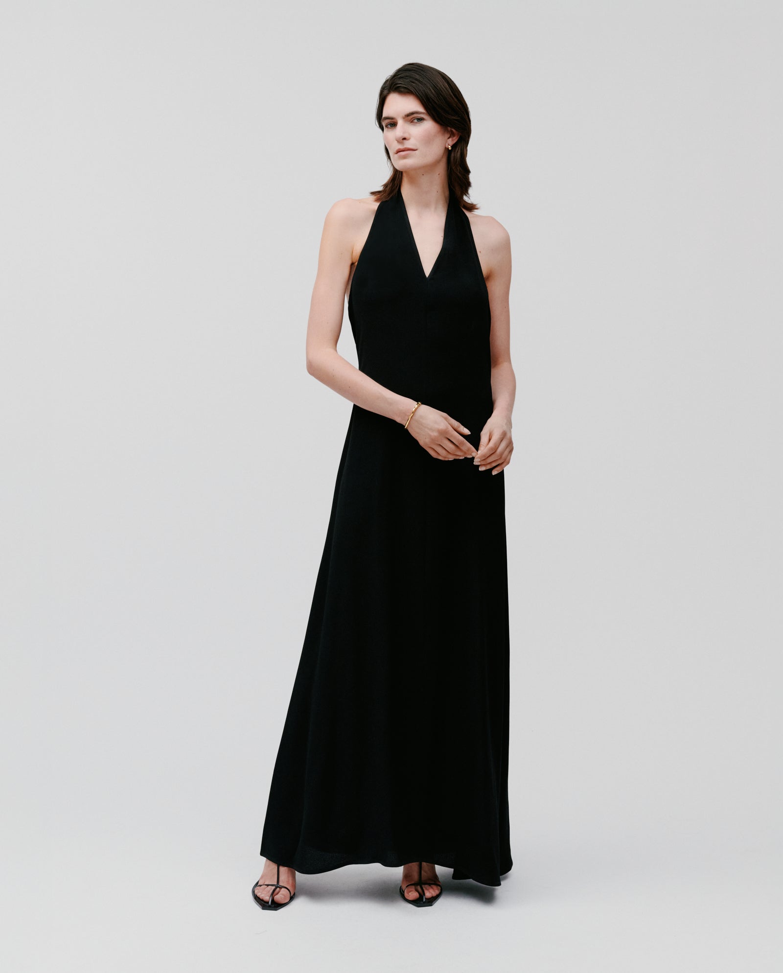 A woman wearing the NESTA Dress by IVY OAK, a long black crepe neckholder dress, paired with black heels, stands against a plain white background.