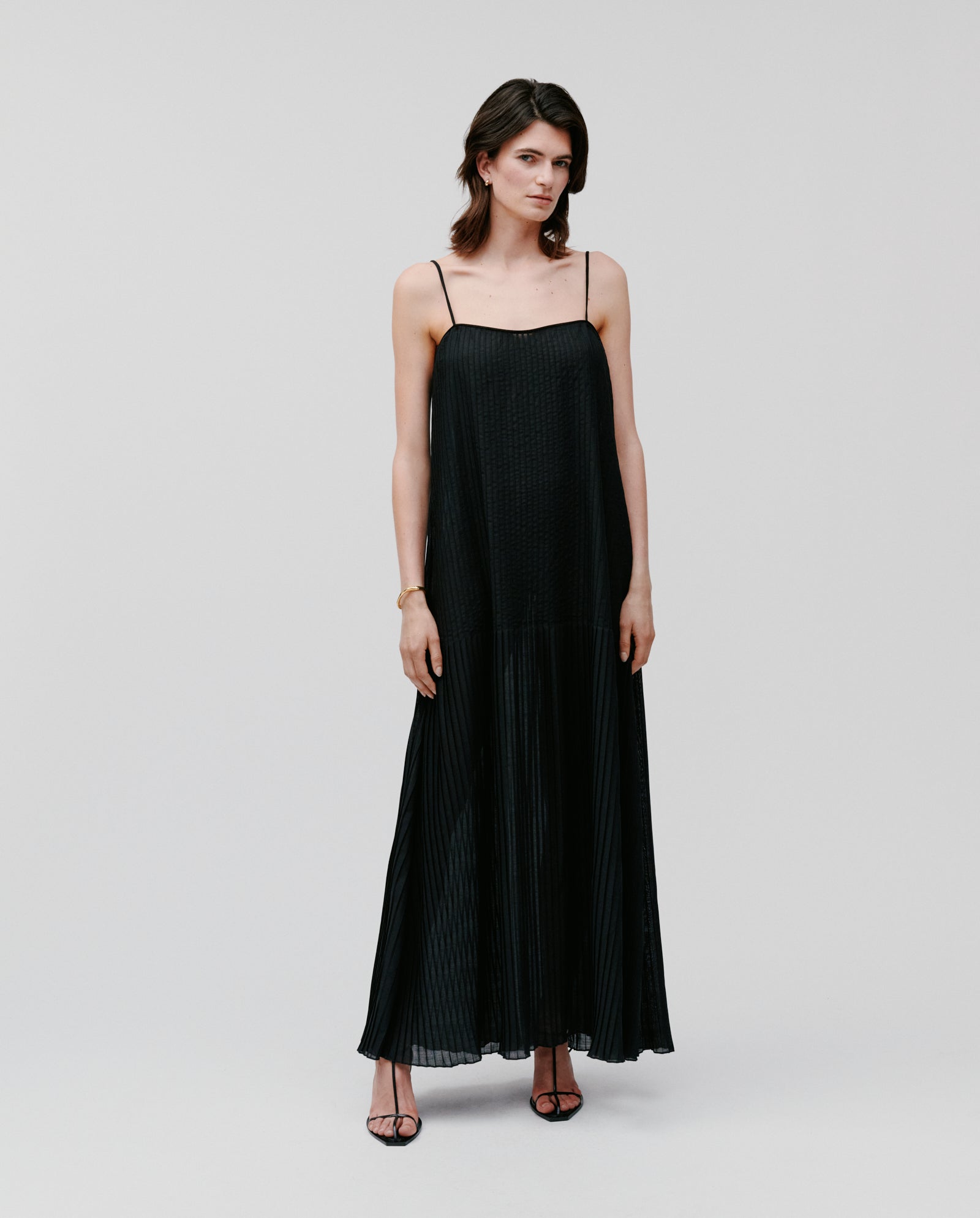 A woman wears the IVY OAK NOE Dress—a long, black plissé gown with thin straps—paired with black heels, standing against a plain background.