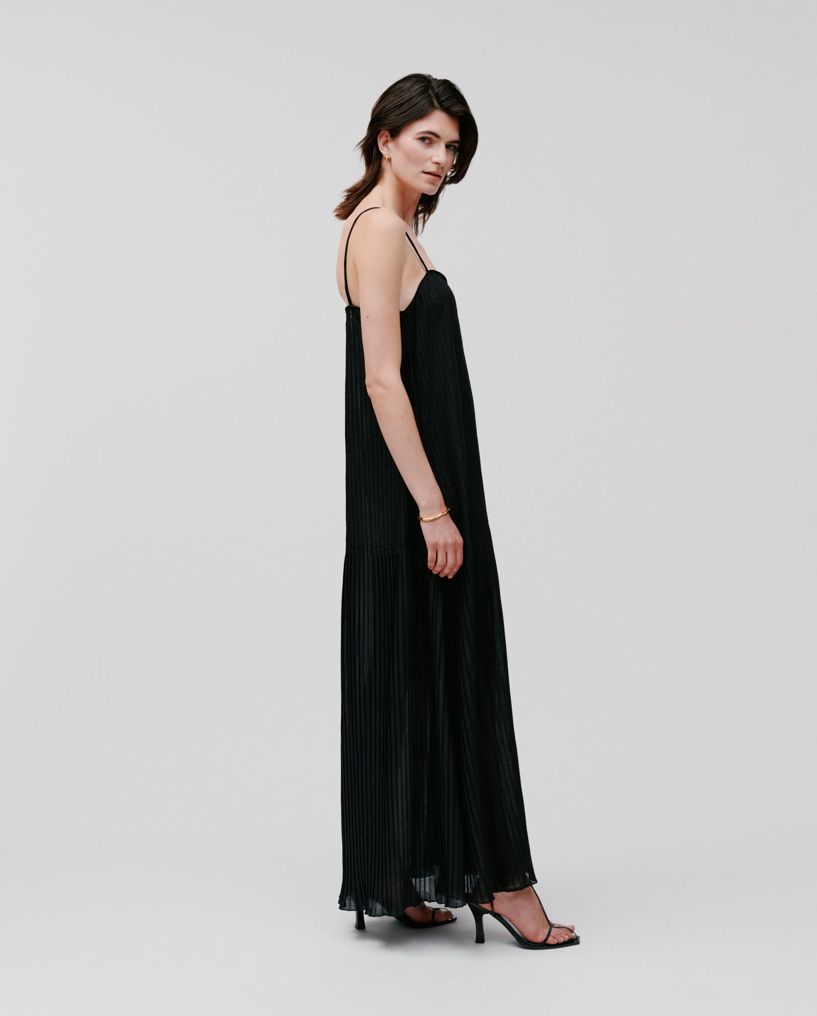 Woman wearing the IVY OAK NOE Dress, a long black plissé fabric design, with black heels, standing sideways against a plain light background.