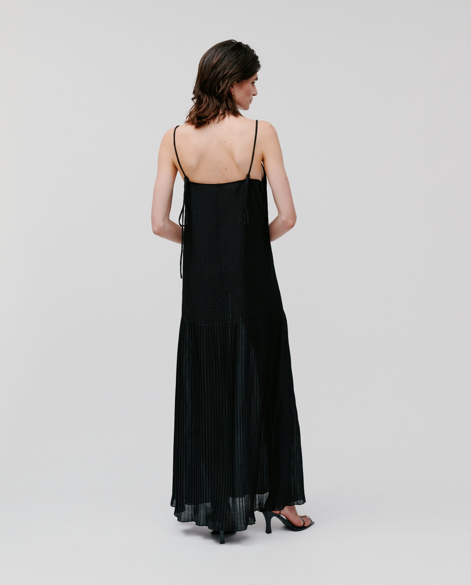 Woman wearing the NOE Dress by IVY OAK, a long black plissé dress with thin straps, standing and facing away.