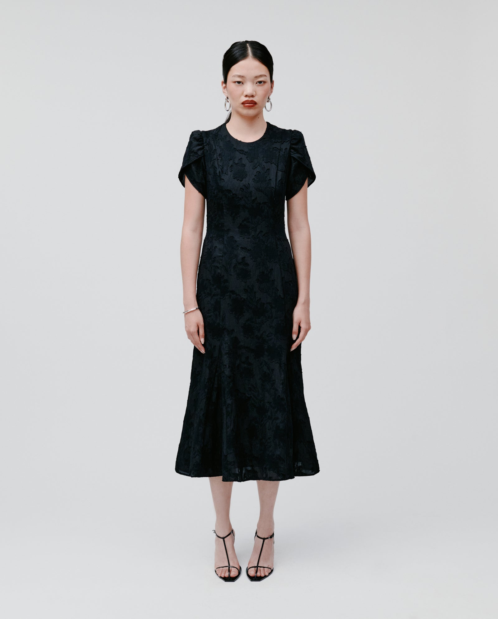 A woman wears the IVY OAK NASRIN Dress, a black midi fil coupé style featuring flower-shaped sleeves and a fitted bodice, paired with strappy heels, standing against a plain background.