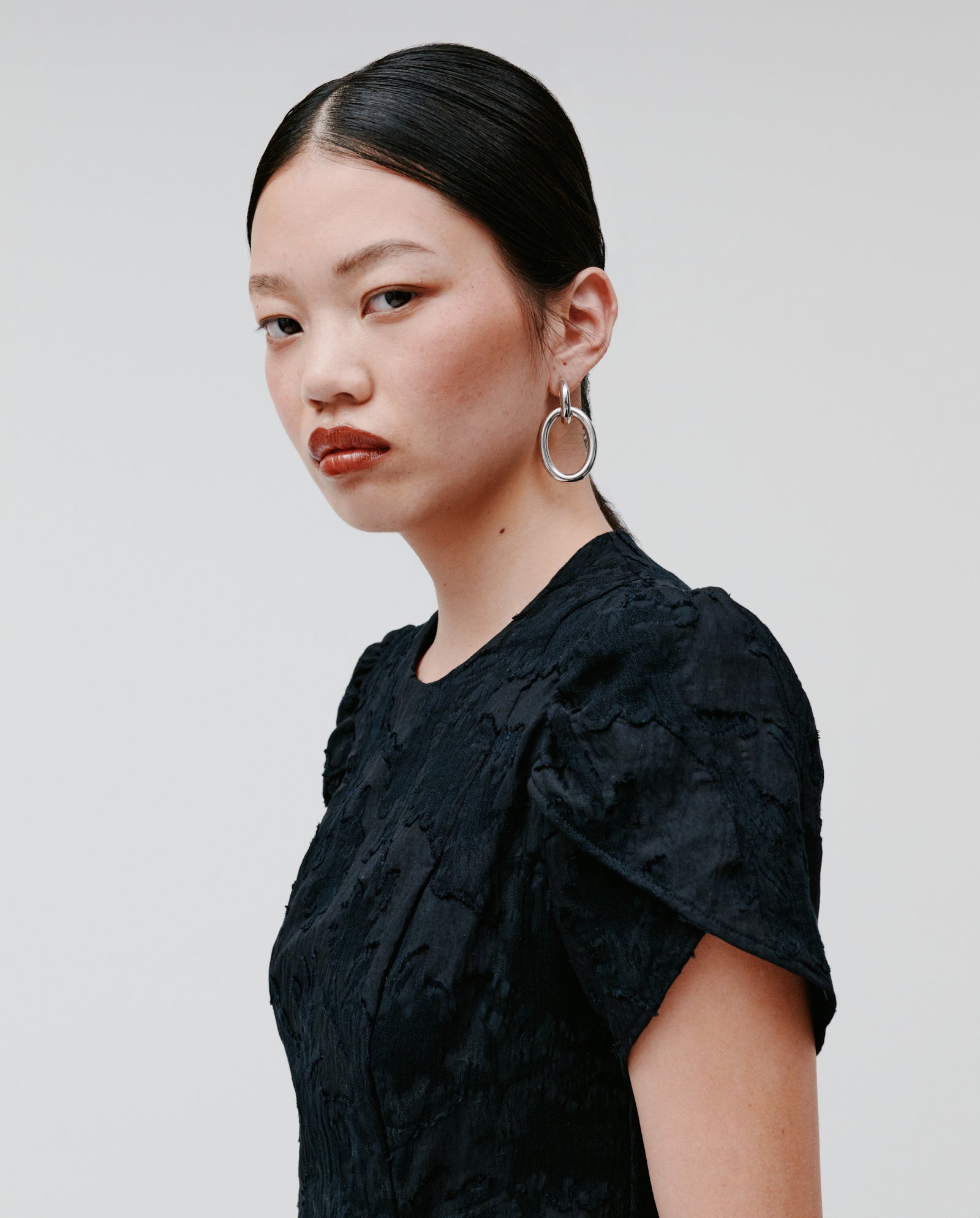 Woman with straight black hair wears the IVY OAK NASRIN Dress, a black midi fil coupé design with a fitted bodice and flower-shaped sleeves, paired with silver hoop earrings, looking at the camera.