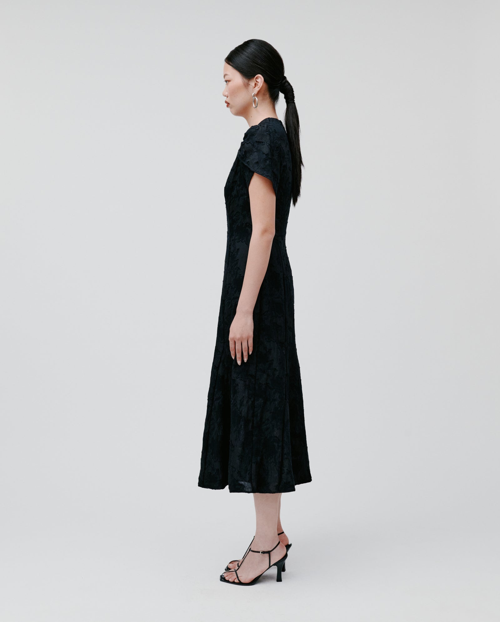 A woman in the IVY OAK NASRIN Dress, featuring a fitted bodice and flower-shaped sleeves, pairs it with heels and poses in profile against a plain light background.