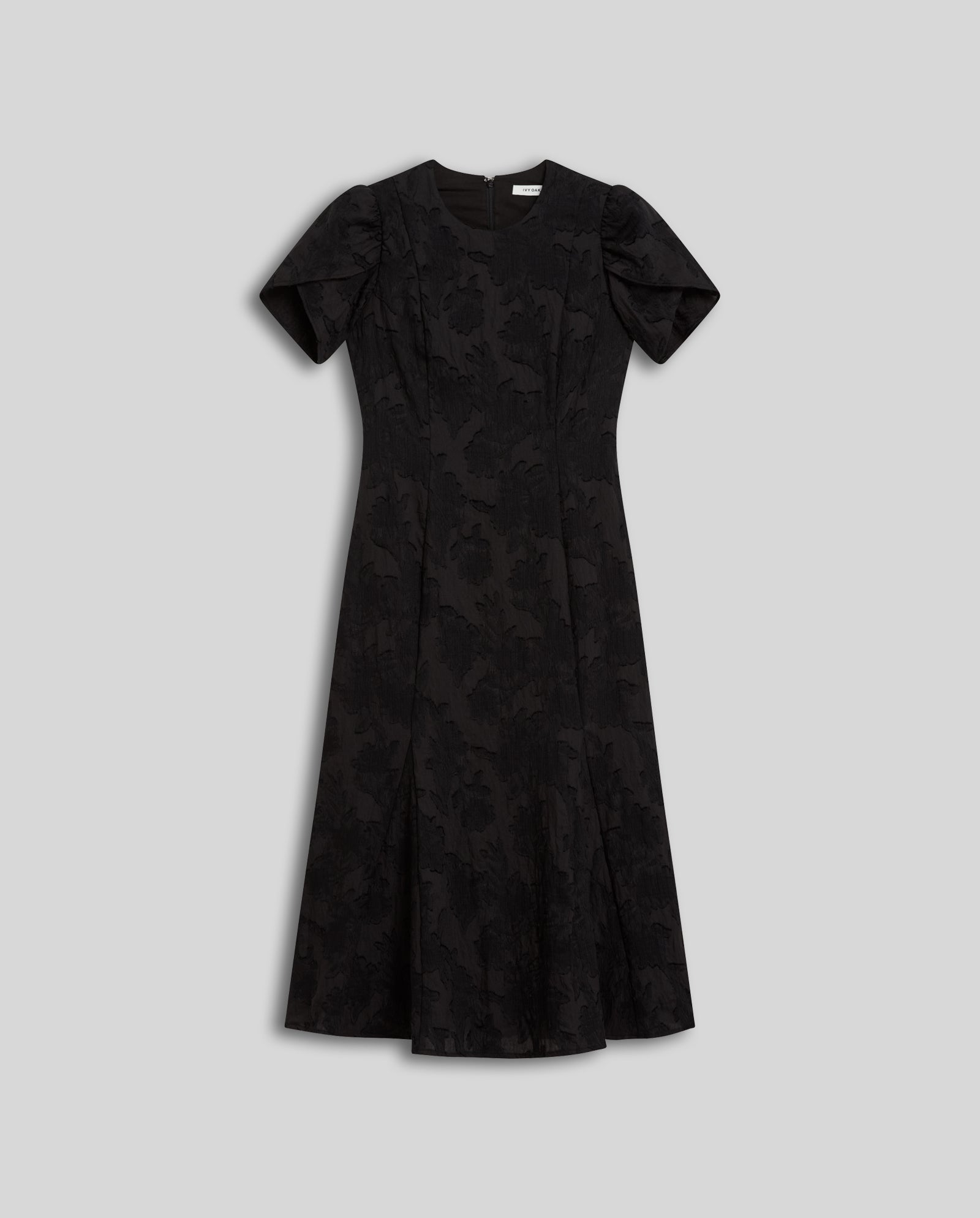 The NASRIN Dress by IVY OAK is a black, short-sleeve midi fil coupé dress with a subtle floral pattern and fitted bodice, shown laid flat on a plain gray background.