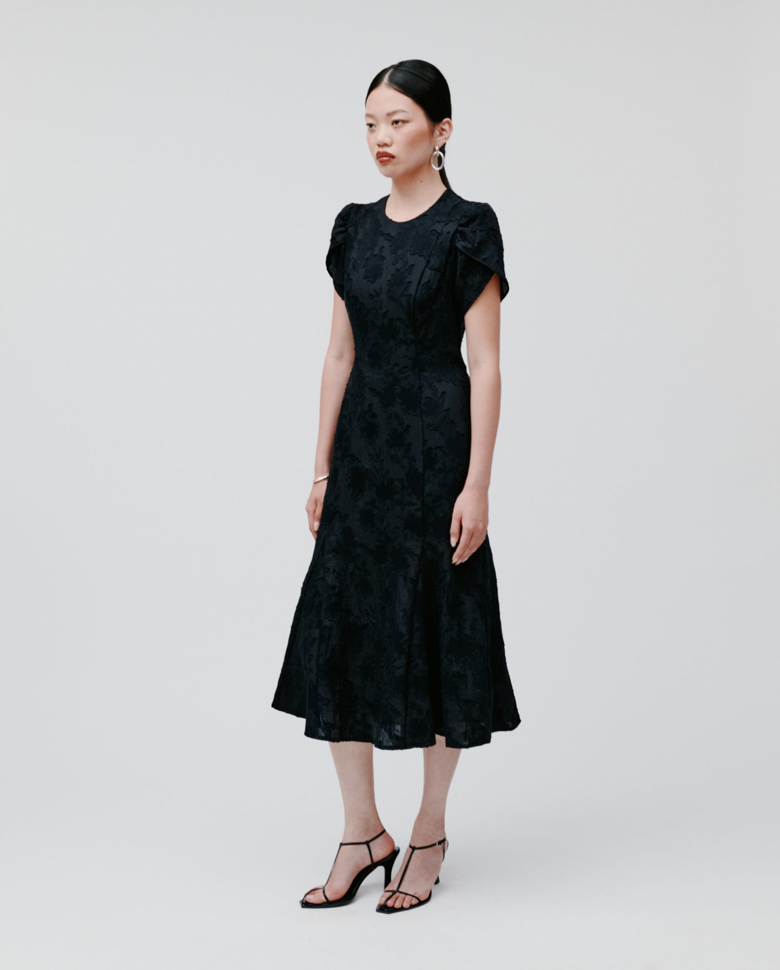 A woman stands against a plain light background wearing the IVY OAK NASRIN black midi fil coupé dress with a fitted bodice, short flower-shaped sleeves, and black heels.