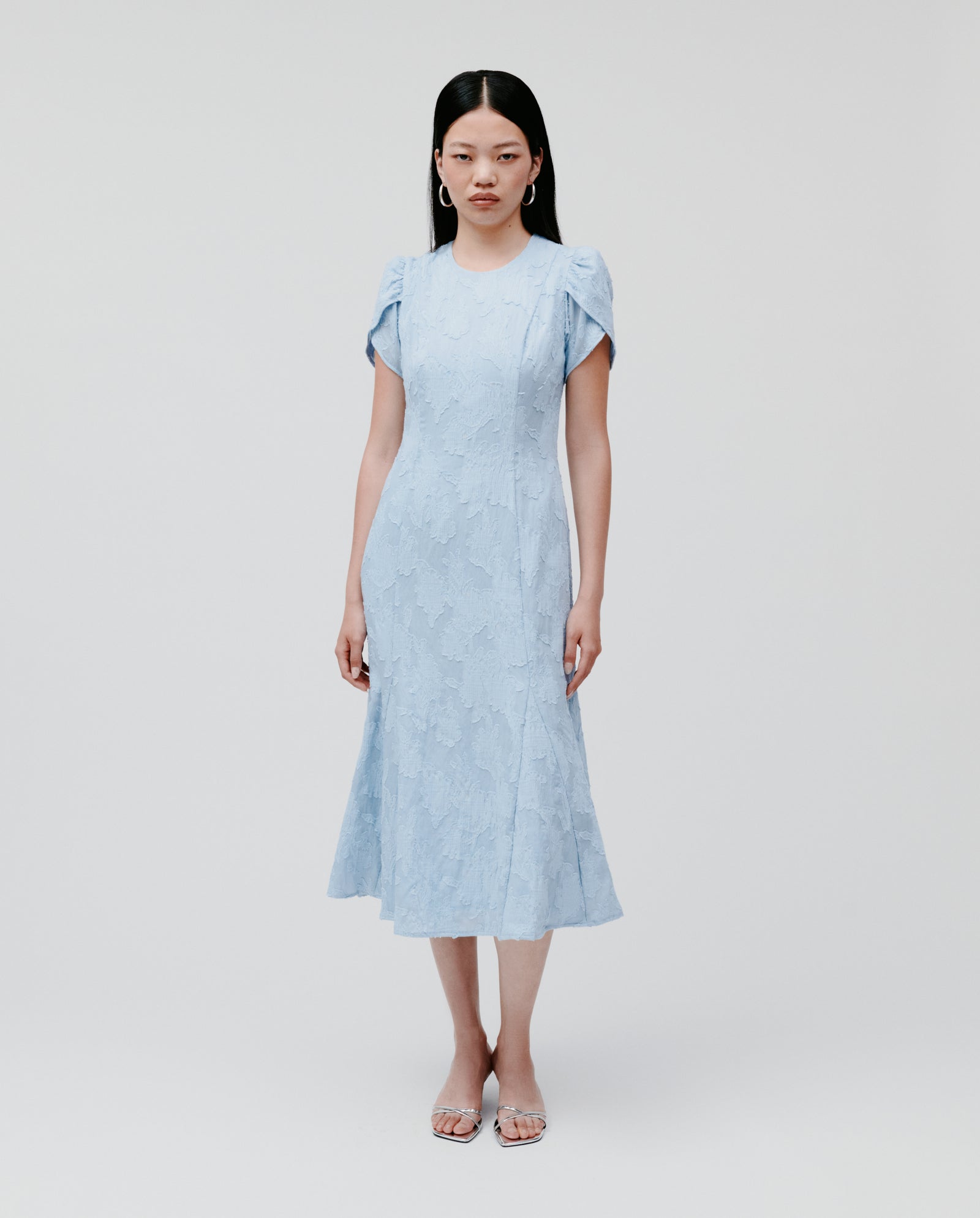 A woman wears the IVY OAK NASRIN Dress, a light blue midi with short sleeves, a fitted bodice, and delicate floral textures, standing against a plain background.