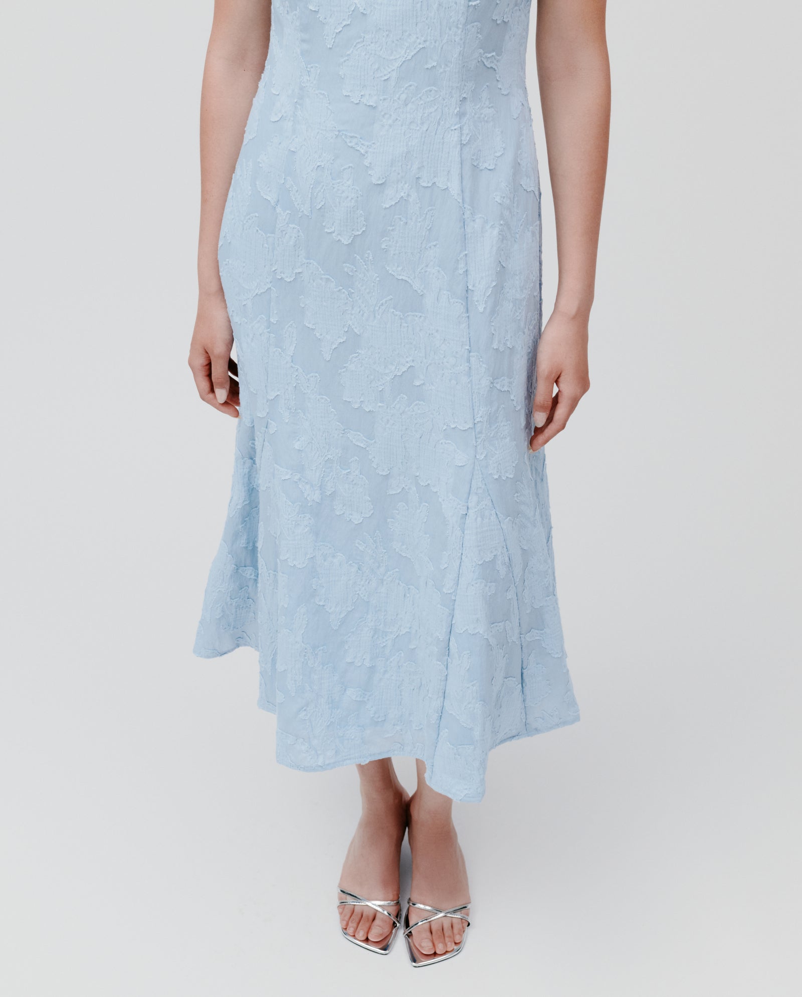 Woman wearing the IVY OAK NASRIN light blue midi dress with floral fil coupé texture and a fitted bodice, paired with silver open-toe heels, standing.