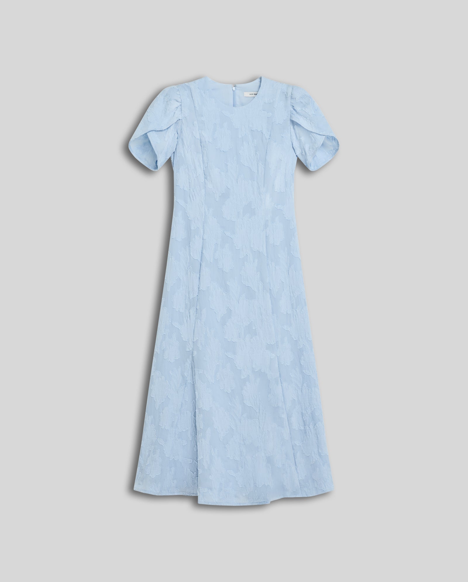 The NASRIN Dress by IVY OAK is a light blue midi fil coupé dress featuring a fitted bodice, subtle floral texture, short sleeves, and an ankle-length hem, shown against a gray background.