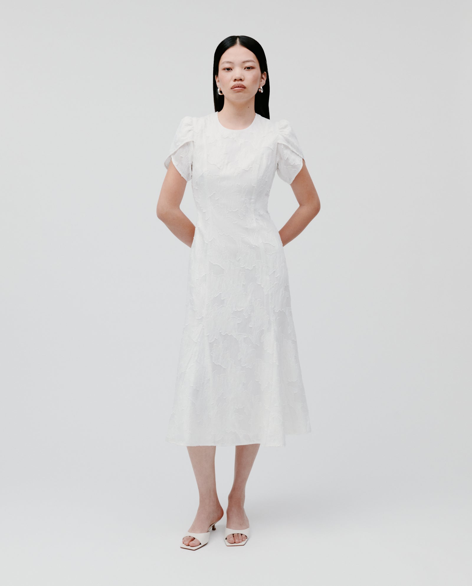Woman in the IVY OAK NASRIN Dress—a white, short-sleeved midi dress with a subtle floral fil coupé pattern and fitted bodice—stands against a plain background.