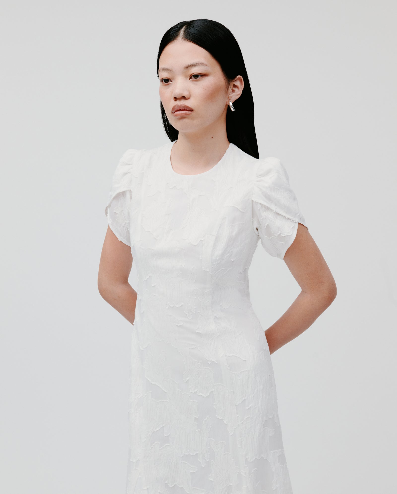 A woman stands with hands behind her back against a plain background, wearing the NASRIN Dress by IVY OAK—a textured white fil coupé midi dress featuring a fitted bodice and short sleeves.