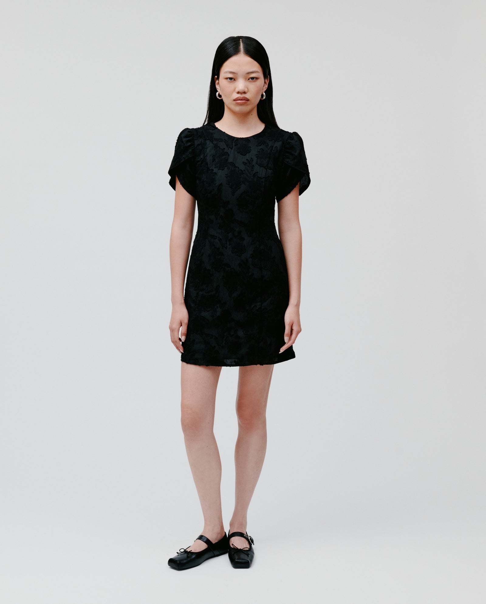 A woman wearing the IVY OAK NERISSA Dress, a black A-line silhouette with short sleeves, pairs it with matching black shoes against a plain white background.
