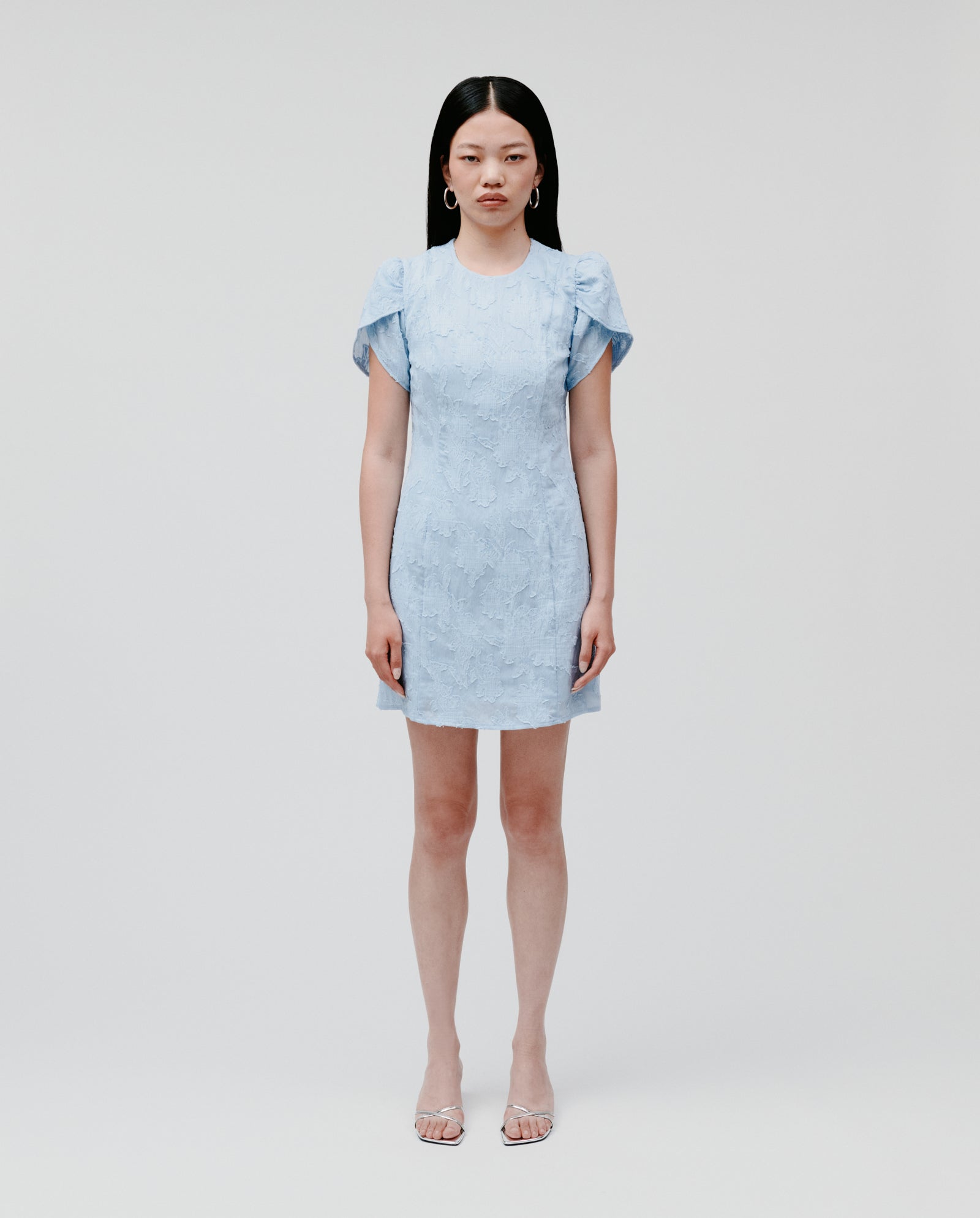A woman poses against a plain background wearing the IVY OAK NERISSA Dress, a light blue fil coupé mini dress with an A-line silhouette, paired with clear high-heeled sandals.