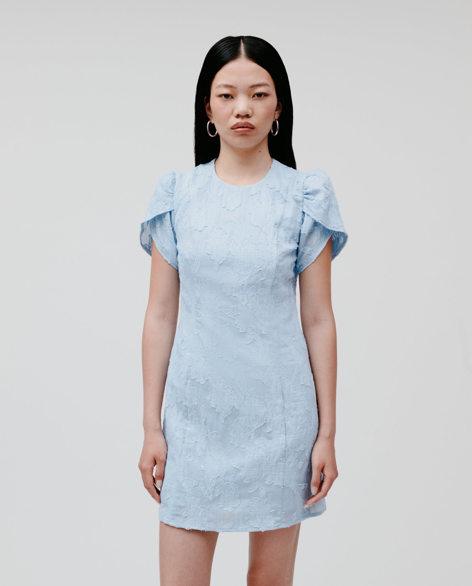 Woman in the IVY OAK NERISSA Dress—a textured light blue fil coupé mini dress featuring short ruffled sleeves and an A-line silhouette—stands against a plain background.