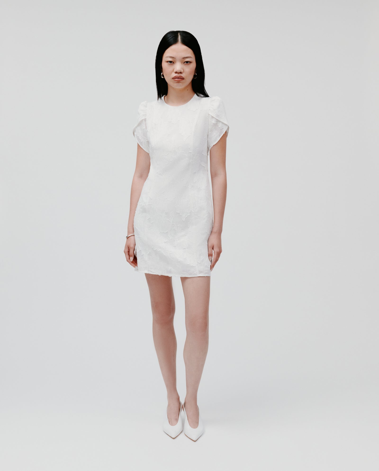 Woman wearing the IVY OAK NERISSA white fil coupé mini dress with floral sleeves and white heels, standing against a plain white background.