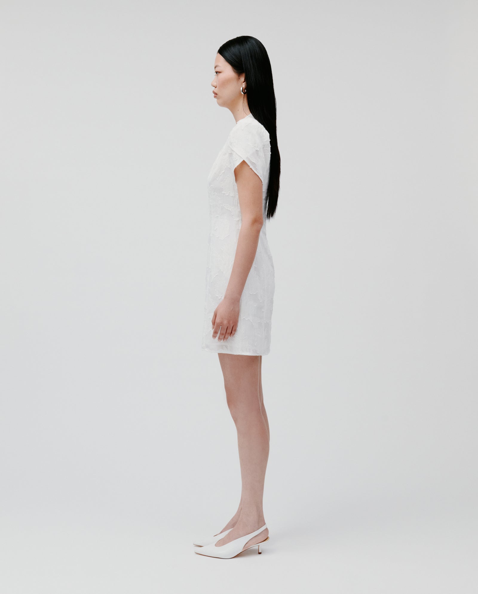 A woman stands in profile against a white background wearing the IVY OAK NERISSA Dress, a fil coupé mini dress with floral sleeves, paired with heels.