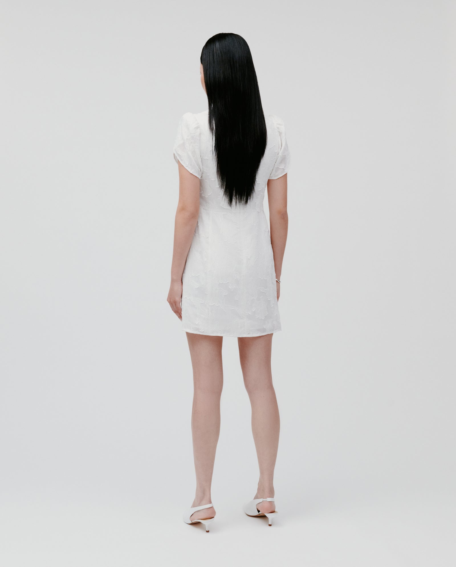 A woman with long dark hair wears the IVY OAK NERISSA Dress and heels, standing facing away in a minimal studio setting.